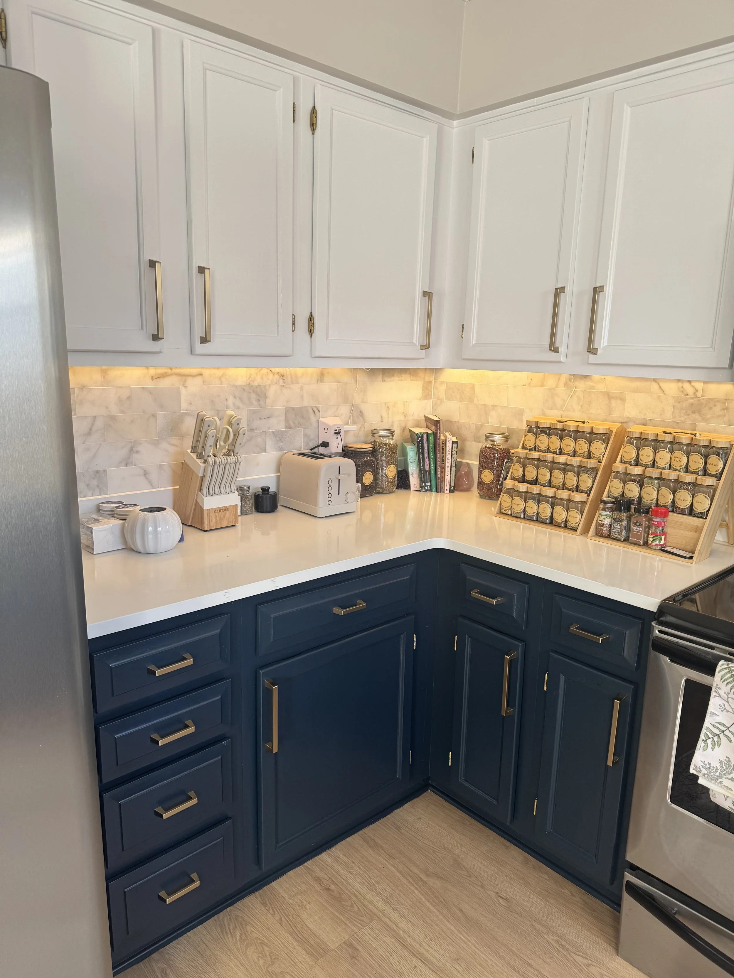 Kitchen corner with white upper cabinets and navy lower cabinets, marble backsplash, countertop with spice jars, knife block, toaster, books, and kitchen tools.