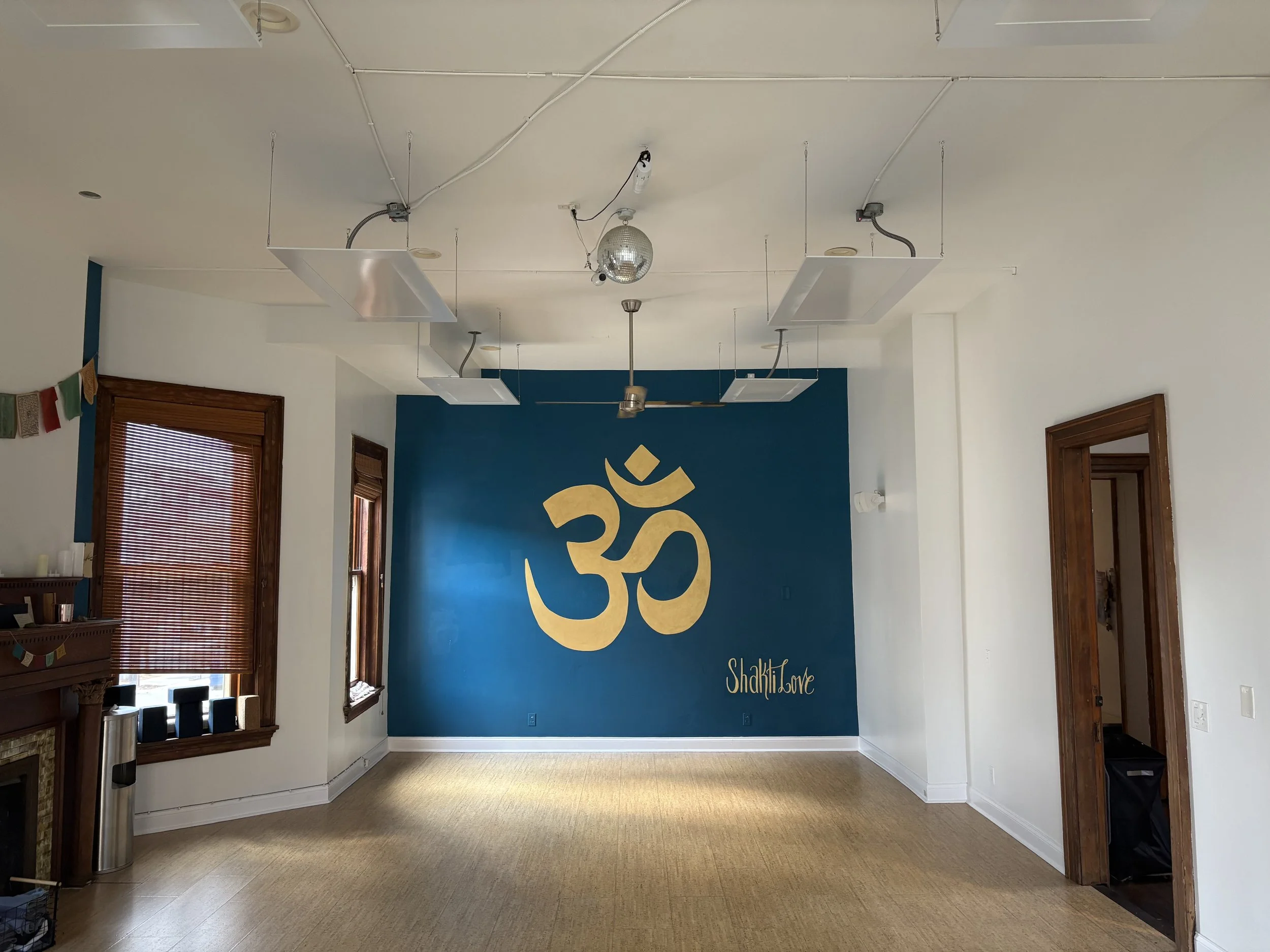 Empty room with white walls and a blue accent wall featuring a large gold 'Om' symbol and the words 'Shakti Love' in gold text. Two windows with wooden frames and blinds on the left, and a wooden door on the right. Ceiling has hanging metallic panels and a disco ball. The floor is light-colored wood or laminate.