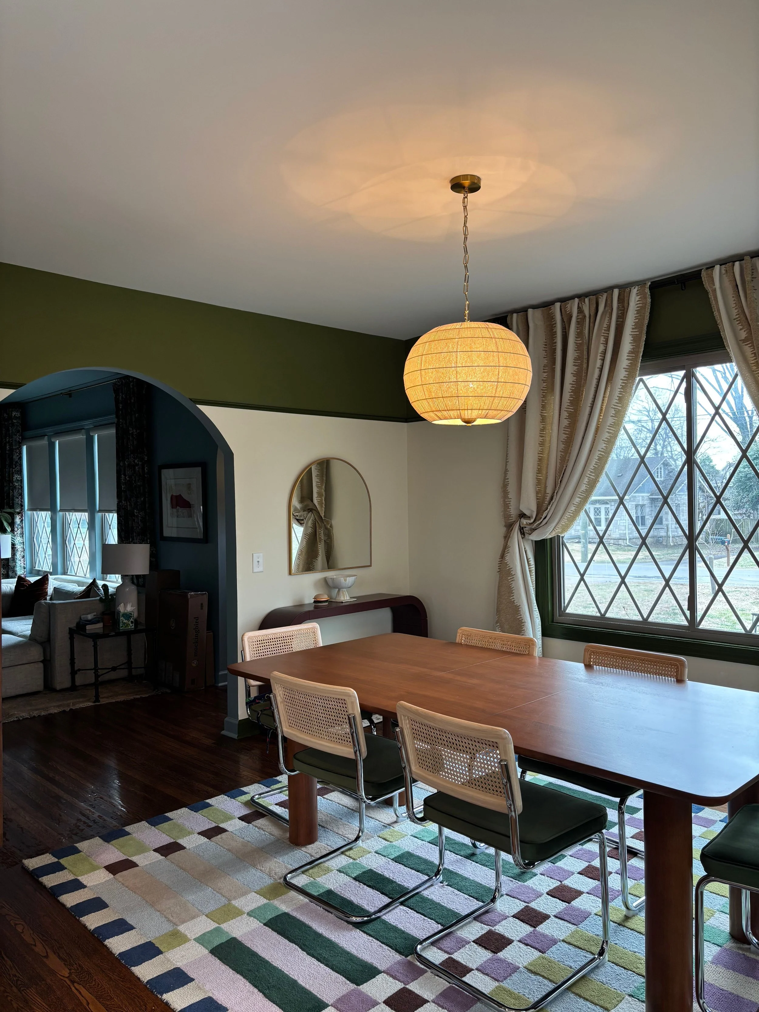 Dining room with a wooden table, six chairs, a hanging paper lantern, a large window with curtains, and a colorful checkered rug.