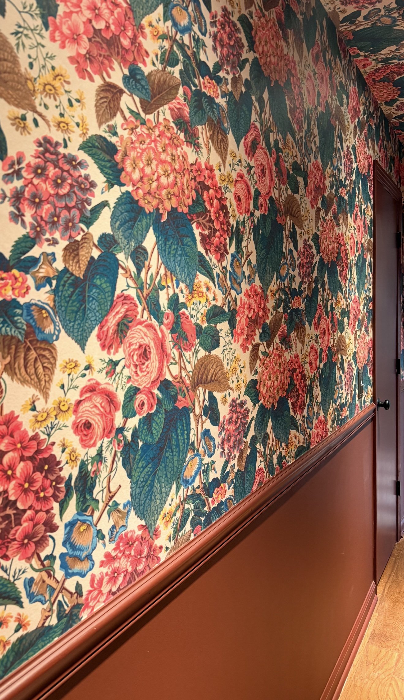 A wall covered with floral wallpaper featuring large pink roses, hydrangeas, and smaller yellow flowers with green and brown leaves. Wall with custom wooden wainscot painted in a reddish-brown color. Divine Savages Wallpaper in Nashville.
