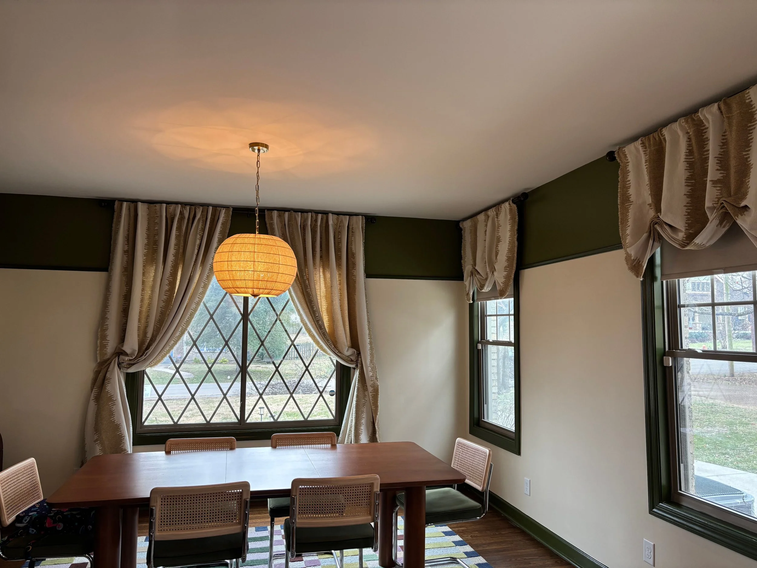 A dining room with a wooden table and six chairs, a window with diamond-shaped panes, and curtains with a striped pattern. A round paper lantern light hangs from the ceiling.