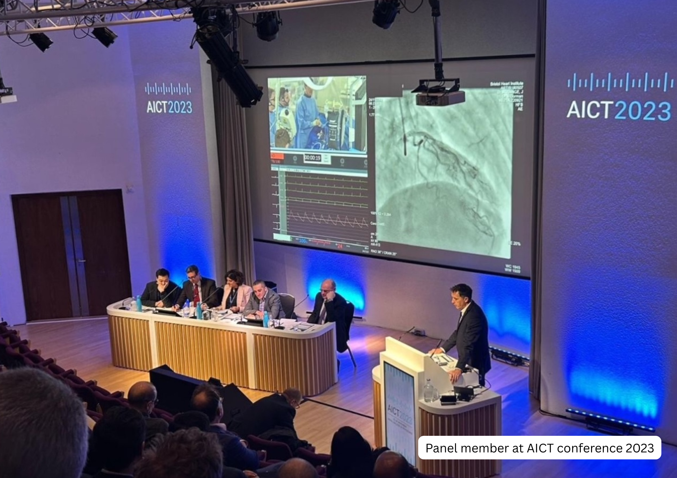 Panel members at AICT conference 2023 with a person at a podium and large screens displaying medical images and data in a conference hall.