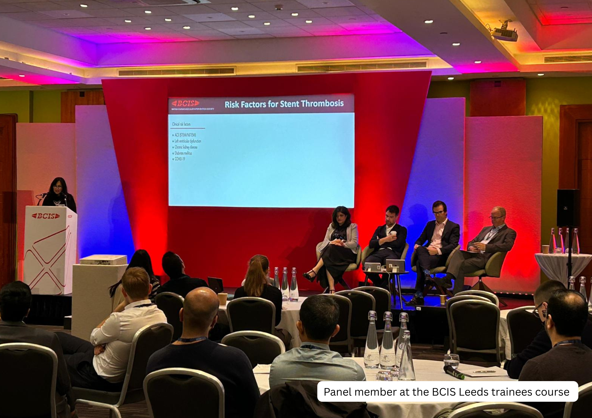 A panel of four people sitting on stage at the BCIS Leeds trainees course, with a presentation slide on risk factors for stent thrombosis displayed behind them, and an audience seated in front.