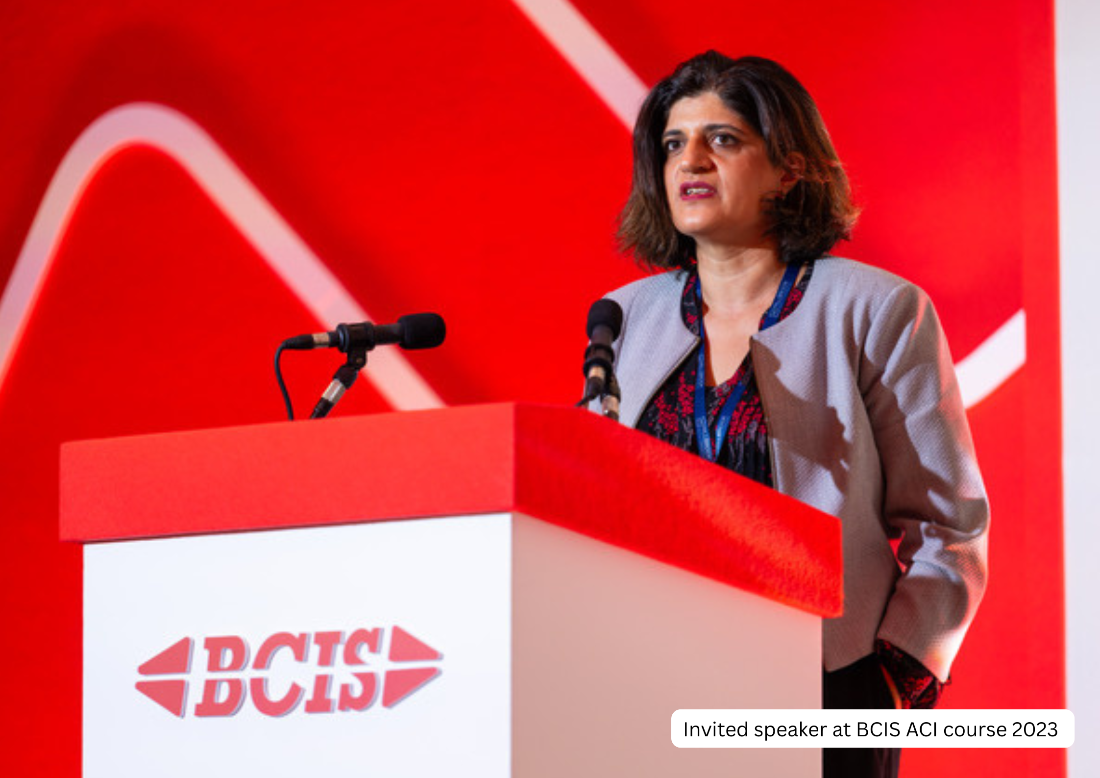 A woman speaking at a podium during a BCIS ACI course 2023, with a red background featuring the BCIS logo and a microphone on the podium.