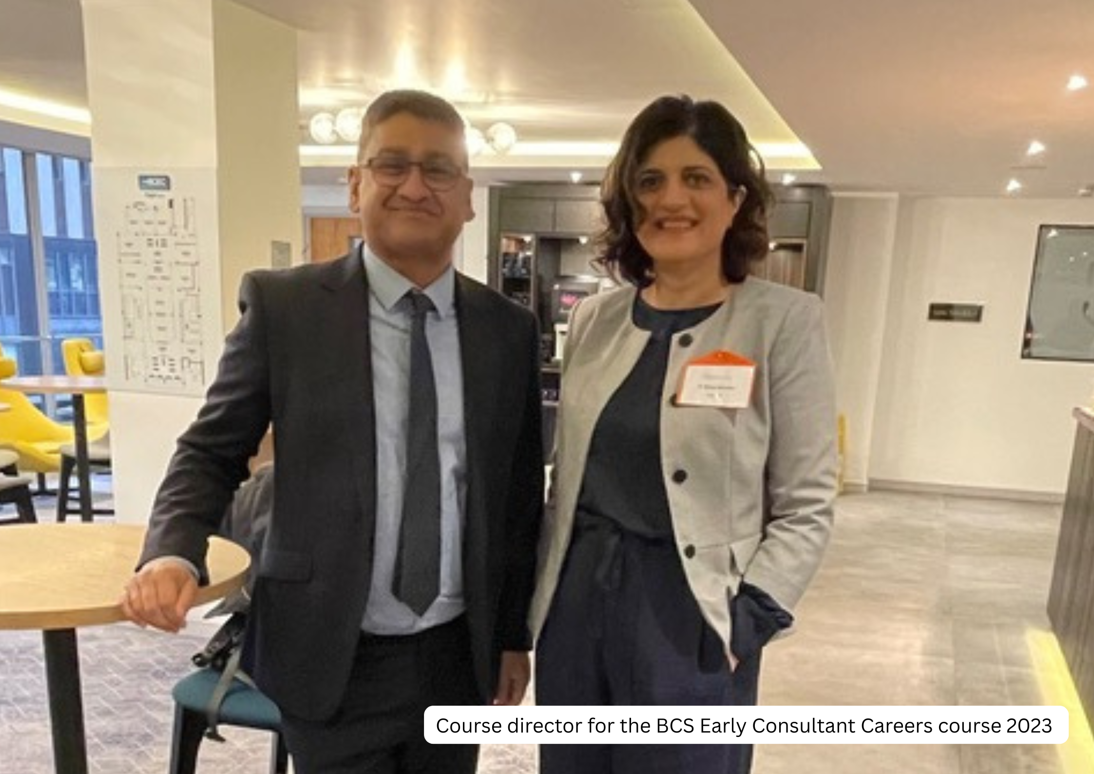 A man and woman standing together in an indoor setting, smiling at the camera. The man is wearing a suit with a tie, and the woman is dressed in a light-colored blazer over a dark top, with a name tag attached to her blazer. The background shows a mo