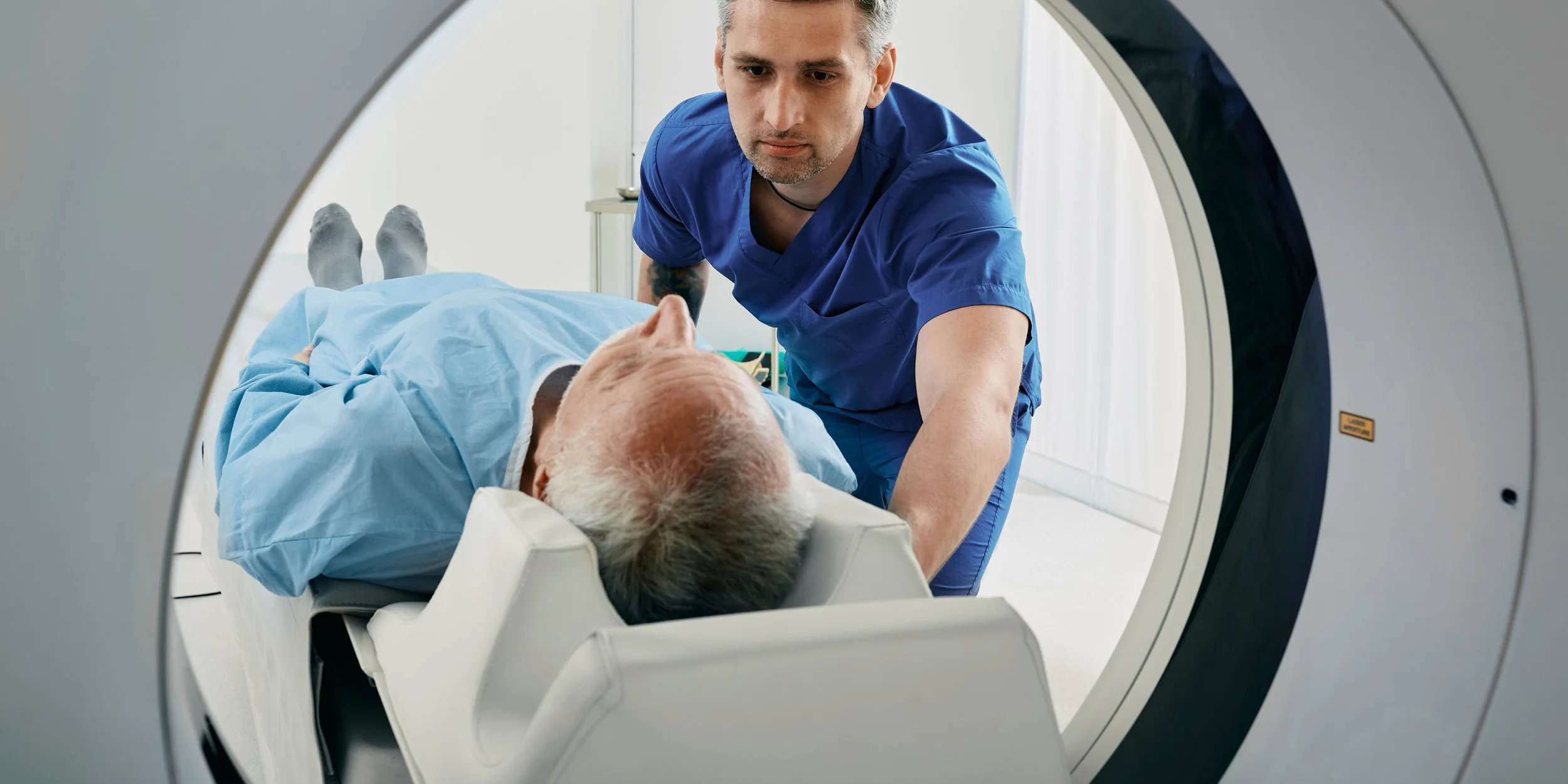 Medical professional in blue scrubs leaning over elderly patient lying on an MRI or CT scanner bed.