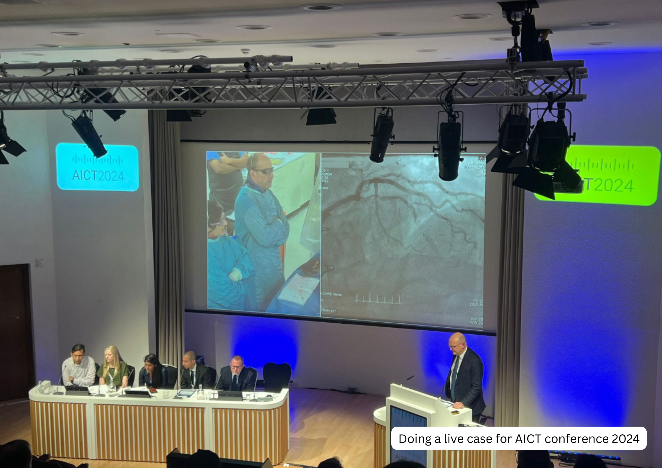 Doctors in surgical scrubs and protective glasses observing an angiogram displayed on a large screen during a medical conference, with a panel of people at a desk and a man at a podium.
