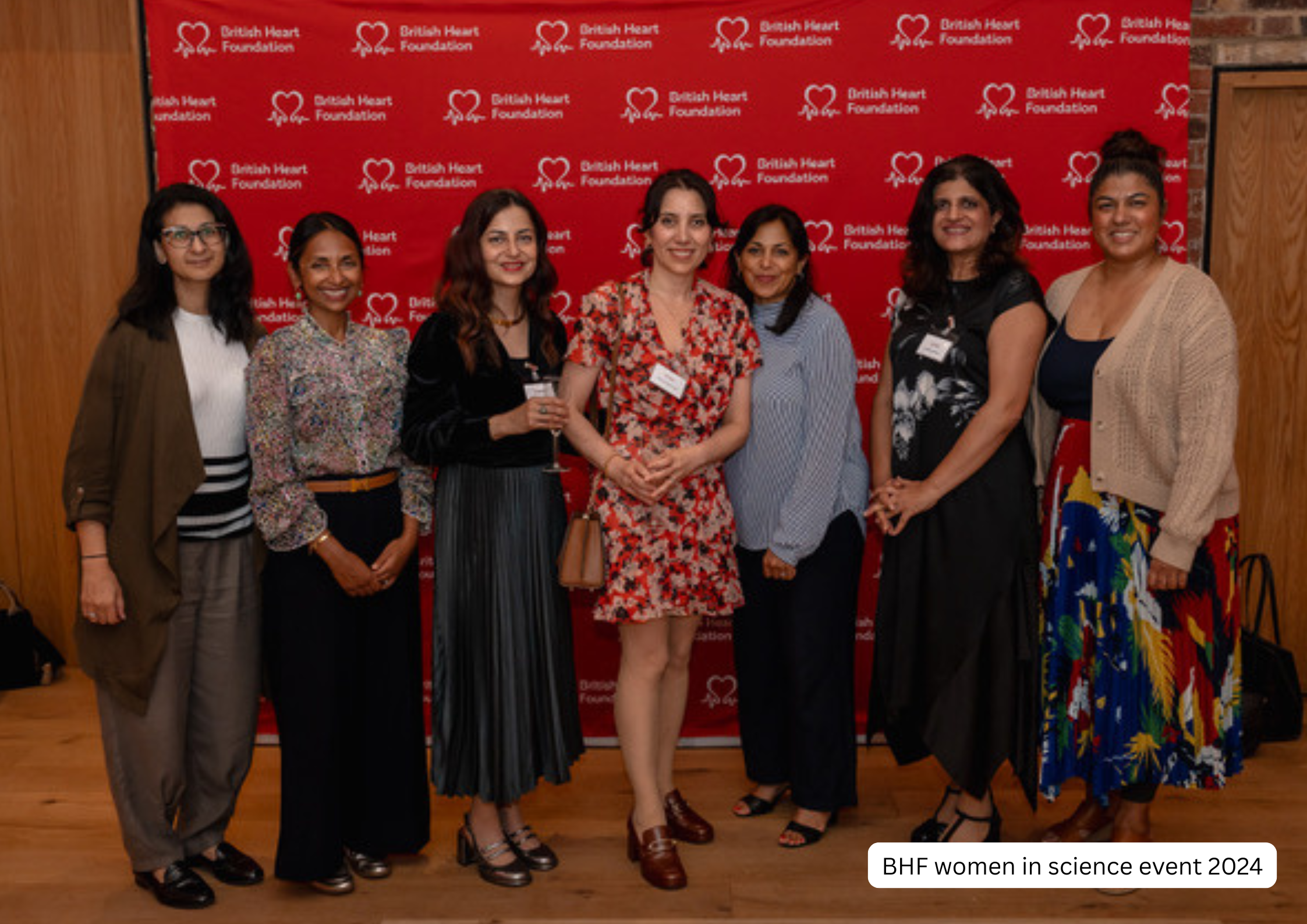 A group of eight women standing together in front of a red backdrop with the British Heart Foundation logo, smiling at the camera at the 2024 women in science event.