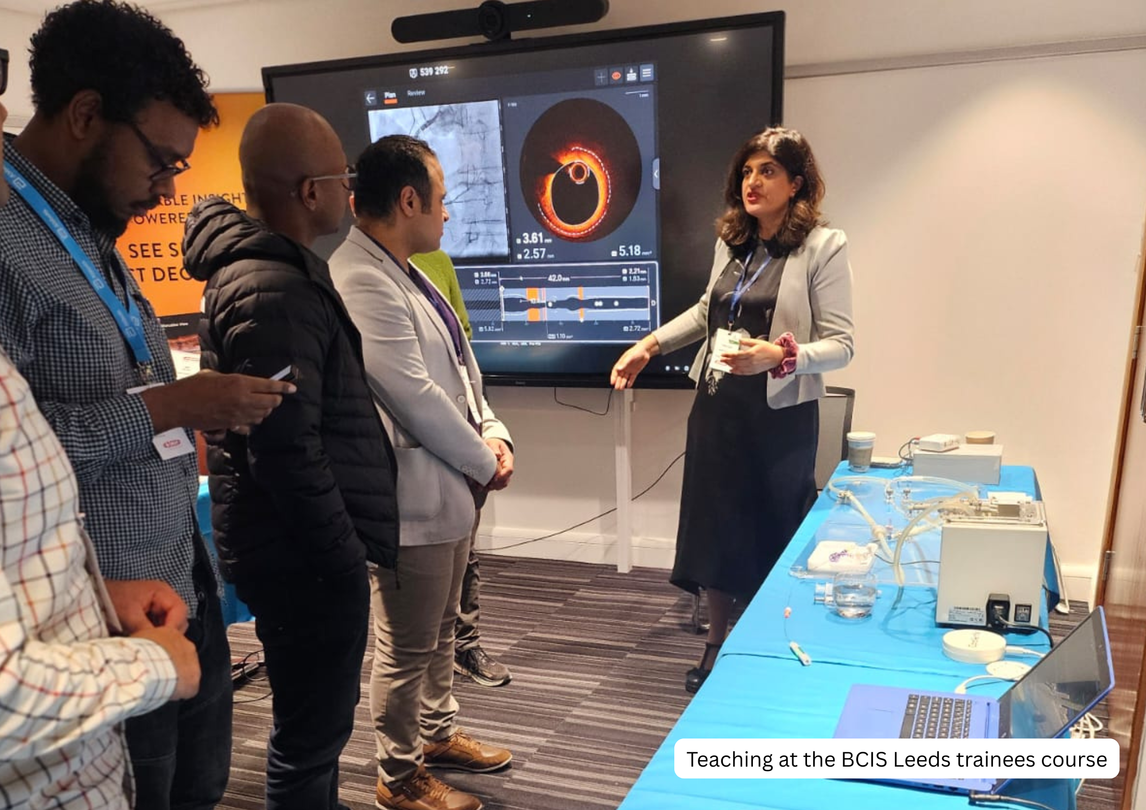 A woman is teaching a group of four people about a scientific topic at a conference, with a large screen behind her displaying scientific diagrams and data related to a medical or biological subject. The group is listening attentively, and there is s