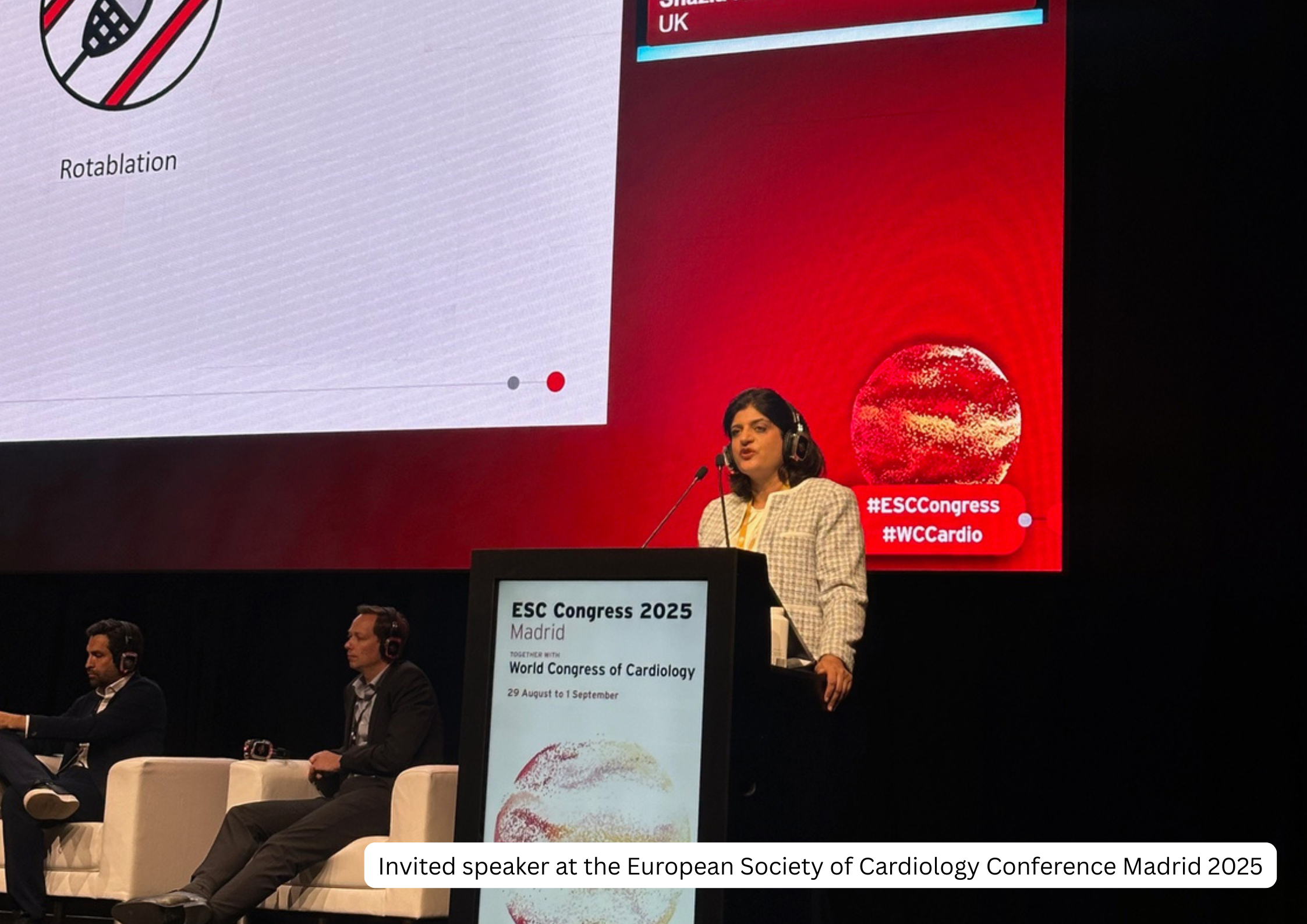 Invited speaker at the European Society of Cardiology Conference Madrid 2025, standing at podium with a large screen displaying a presentation slide and the event details, with two seated panelists in the background.