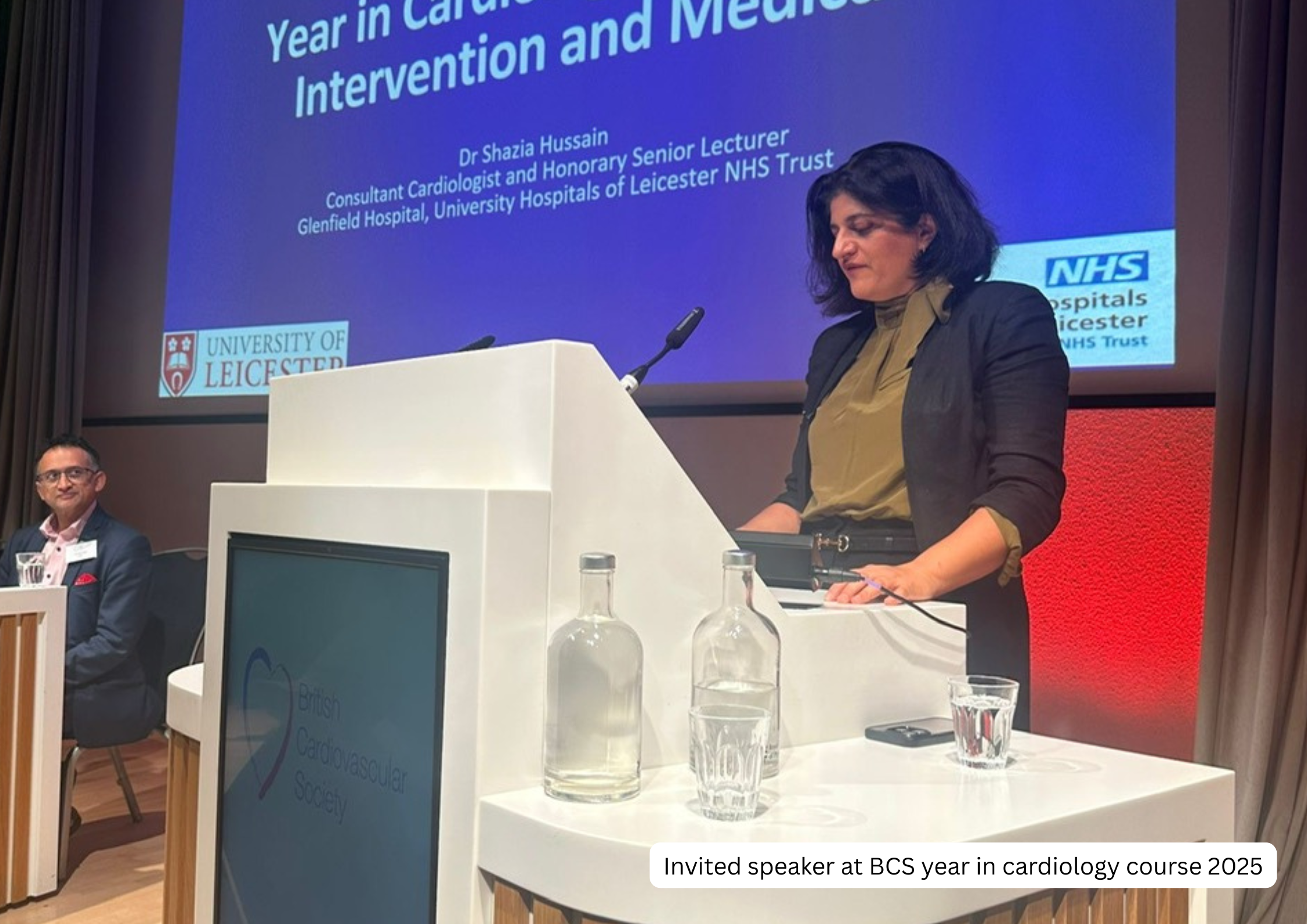 A woman speaking at a podium during a conference, with a large screen behind her displaying the presentation title and speaker information. The screen shows the title "Year in Cardiology: Intervention and Med" and the name Dr. Shazia Hussain, a consu