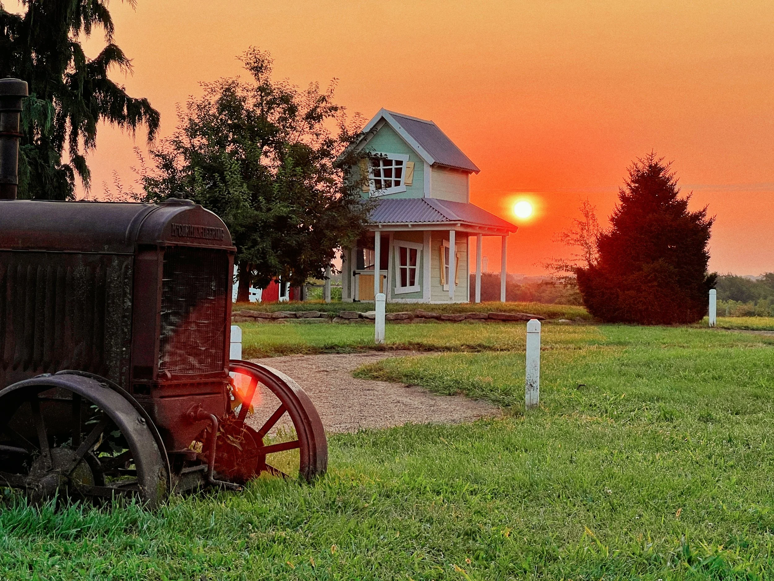 A rural scene during sunset with an old tractor in the foreground, a small house with new windows, and trees in the background.