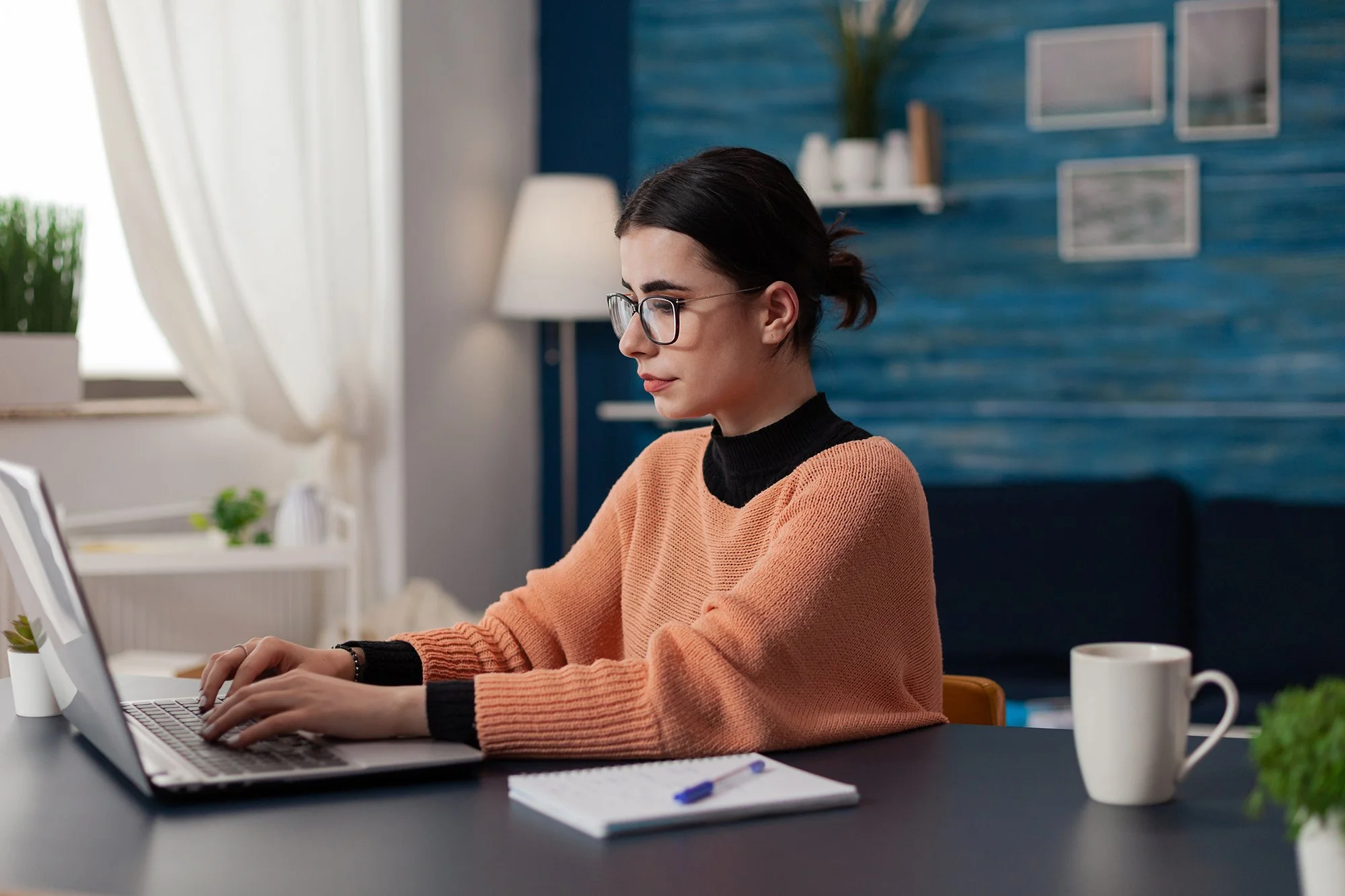 A woman with glasses working on a laptop at a desk, with a notebook and pen, a mug, and plants in a cozy, well-lit home office.