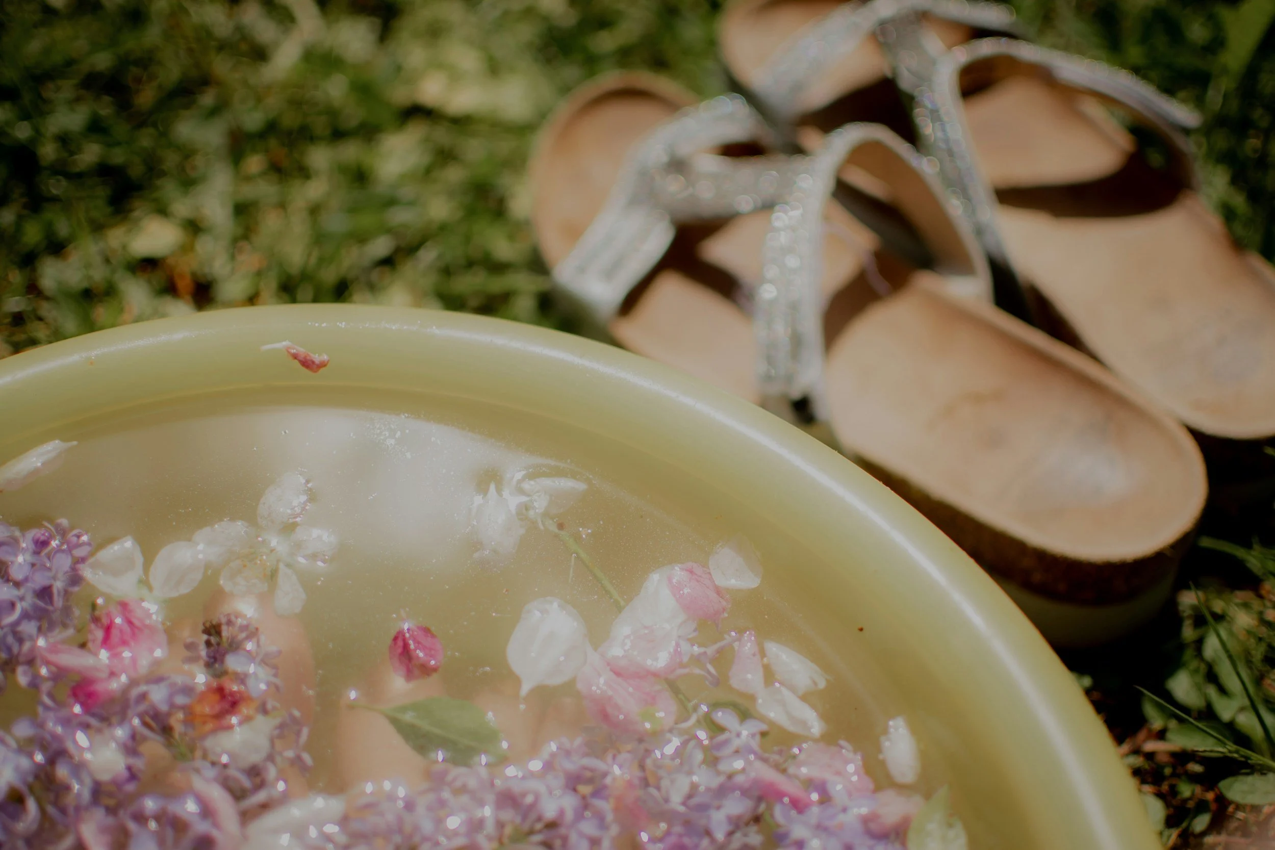 A partially filled yellow bowl with flower petals and small leaves floating on water, with a pair of beige sandals with sparkly straps in the background on grass.