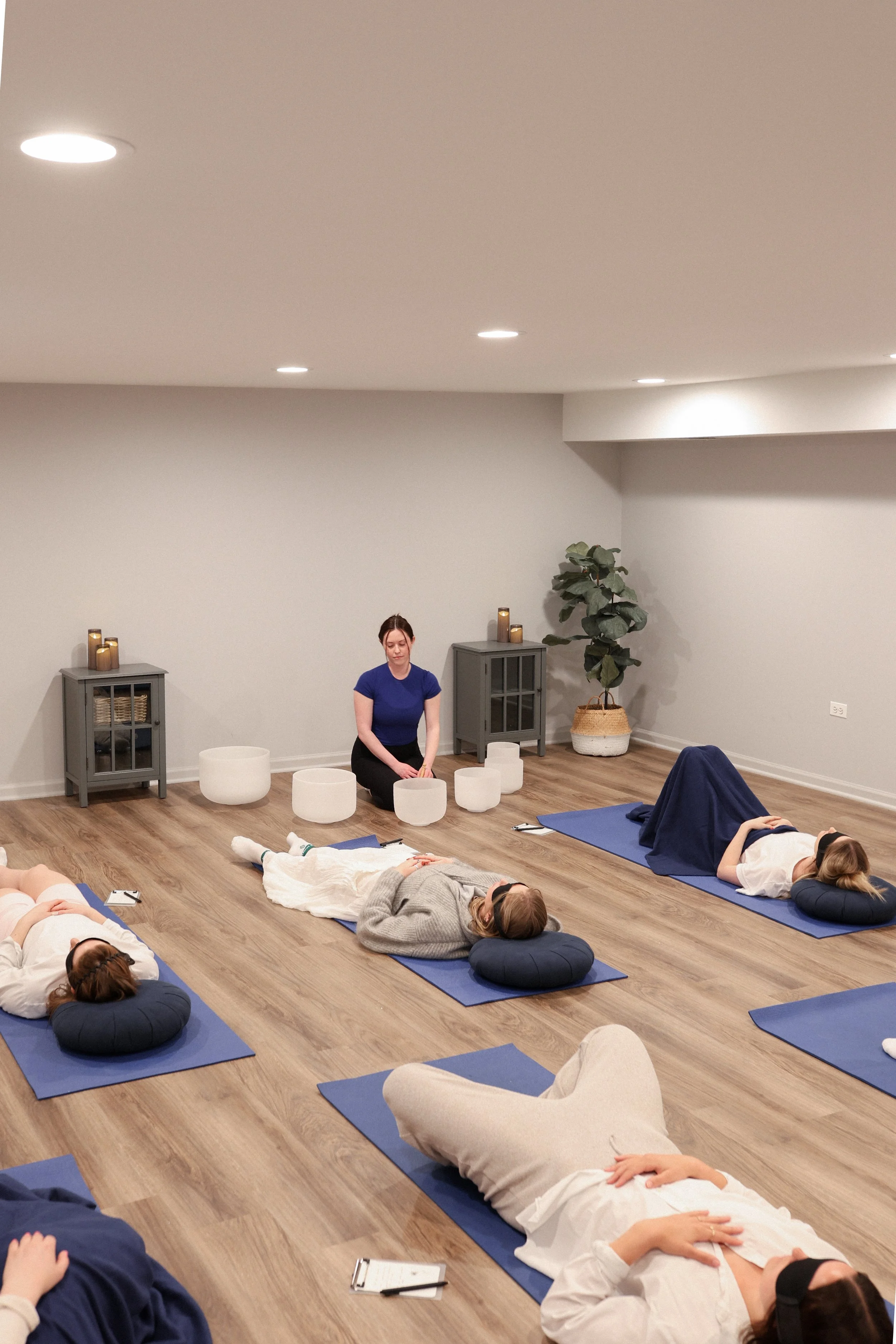 Group of women participating in a mindfulness or meditation class in a minimalist room, lying on mats with pillows, while an instructor guides them.