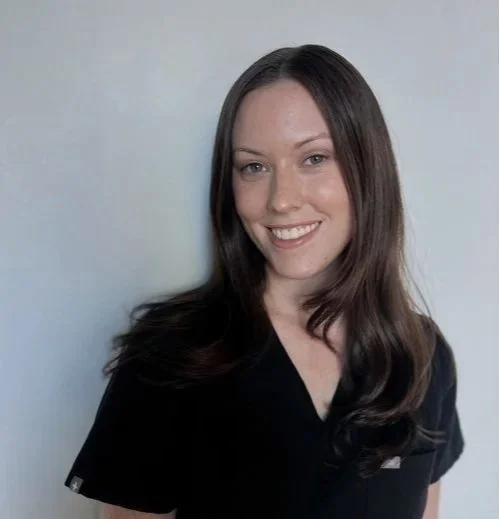 A woman with long dark hair smiling, wearing a black top, standing against a light gray background.
