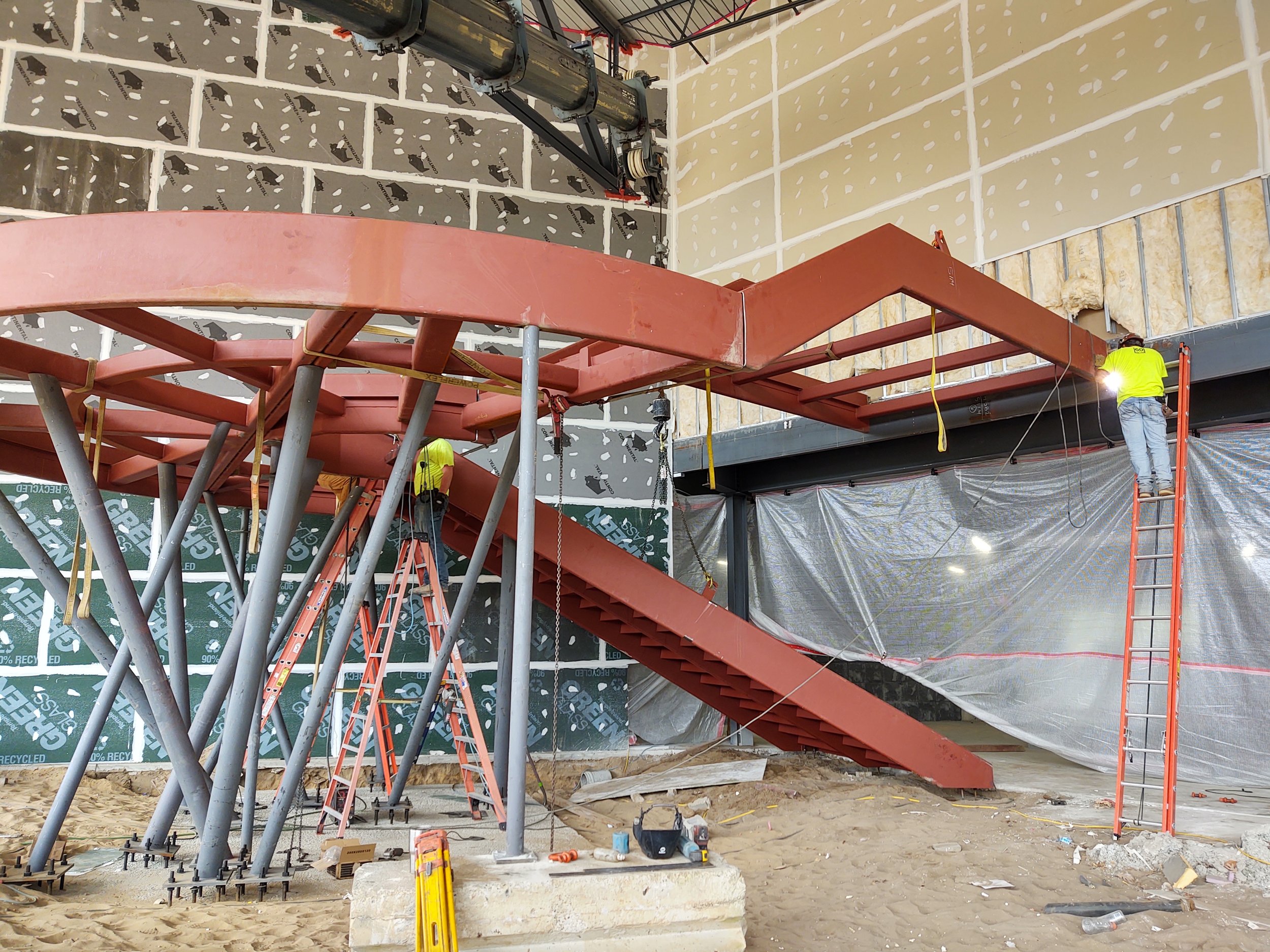Construction site indoors with workers installing a large curved red steel staircase. One worker on a ladder on the right, another worker on the stairs, with tools and materials scattered on the sandy floor.