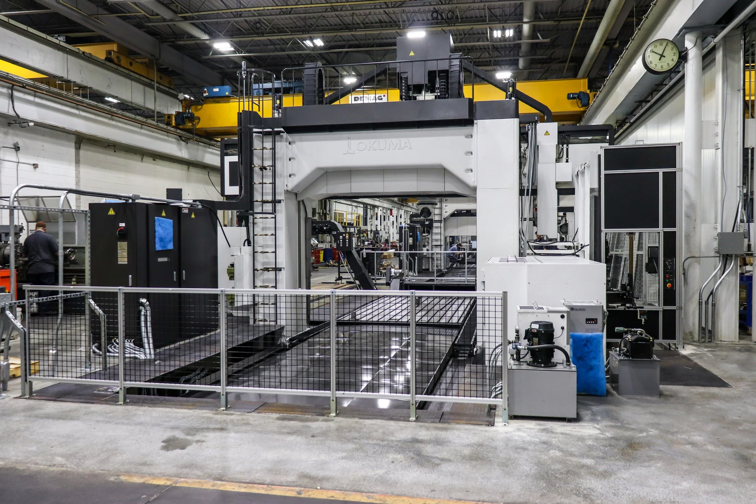 Industrial manufacturing facility with large white CNC machine, metal fenced area, and workers in the background, overhead crane, and industrial clock.