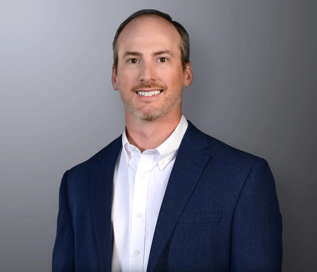 Professional headshot of a man with short hair, light beard, smiling, wearing a white dress shirt and dark blue blazer against a gray background.