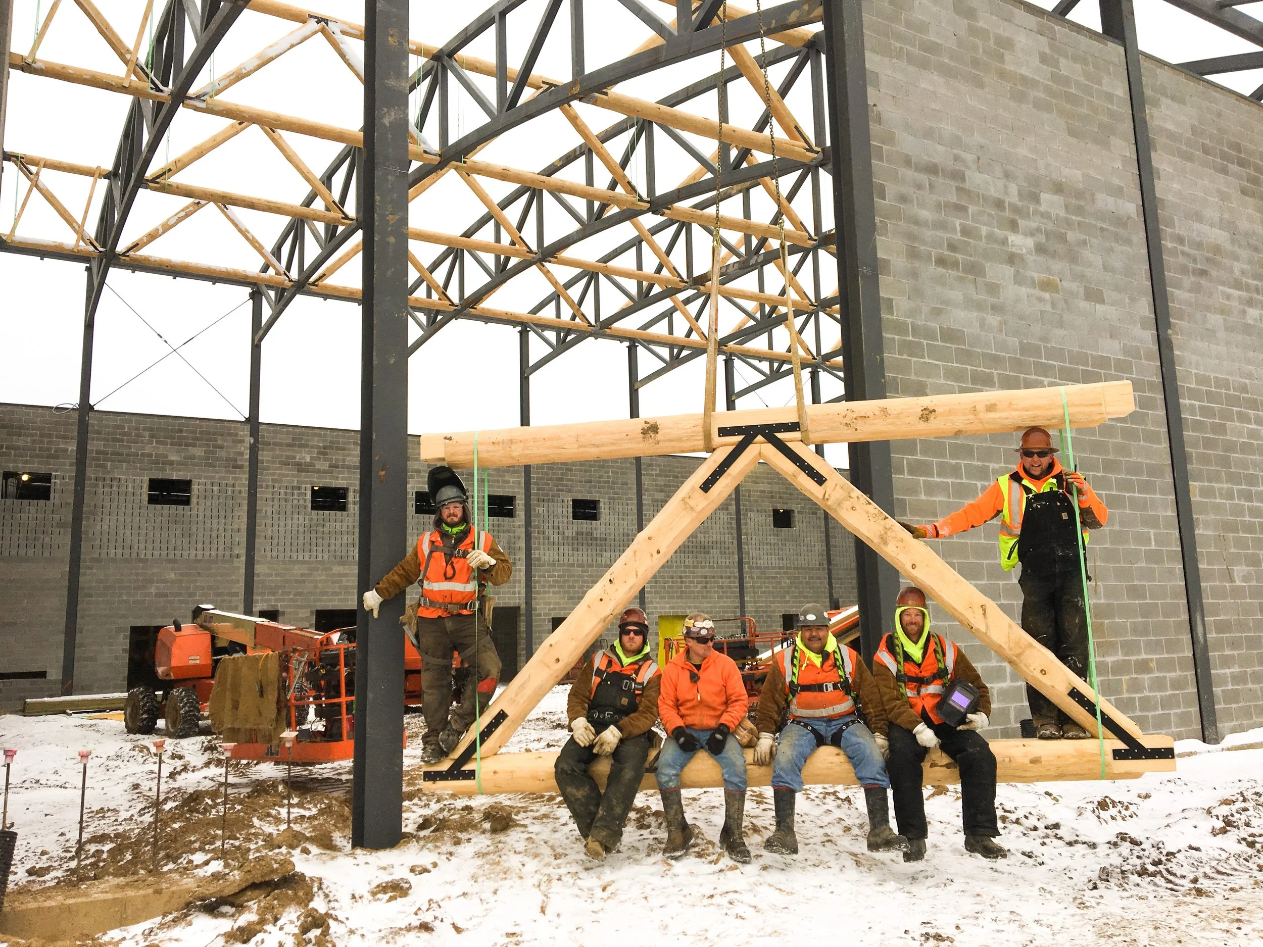 Group of construction workers posing in front of a building under construction, with some sitting and some standing on wooden beams, on a snowy ground.