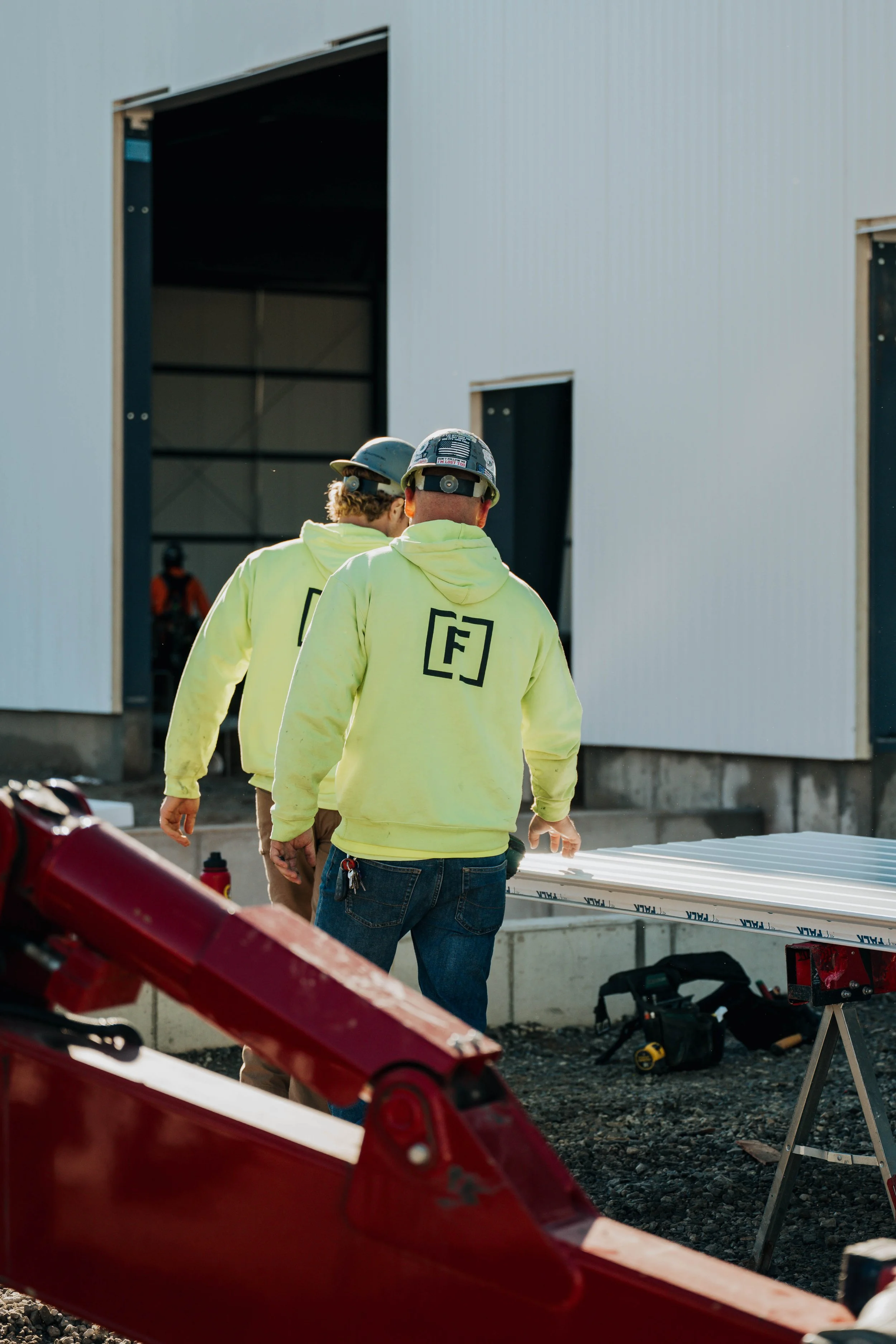 Two FCC Construction crew members in branded hi-vis shirts walk toward a steel building on the jobsite.