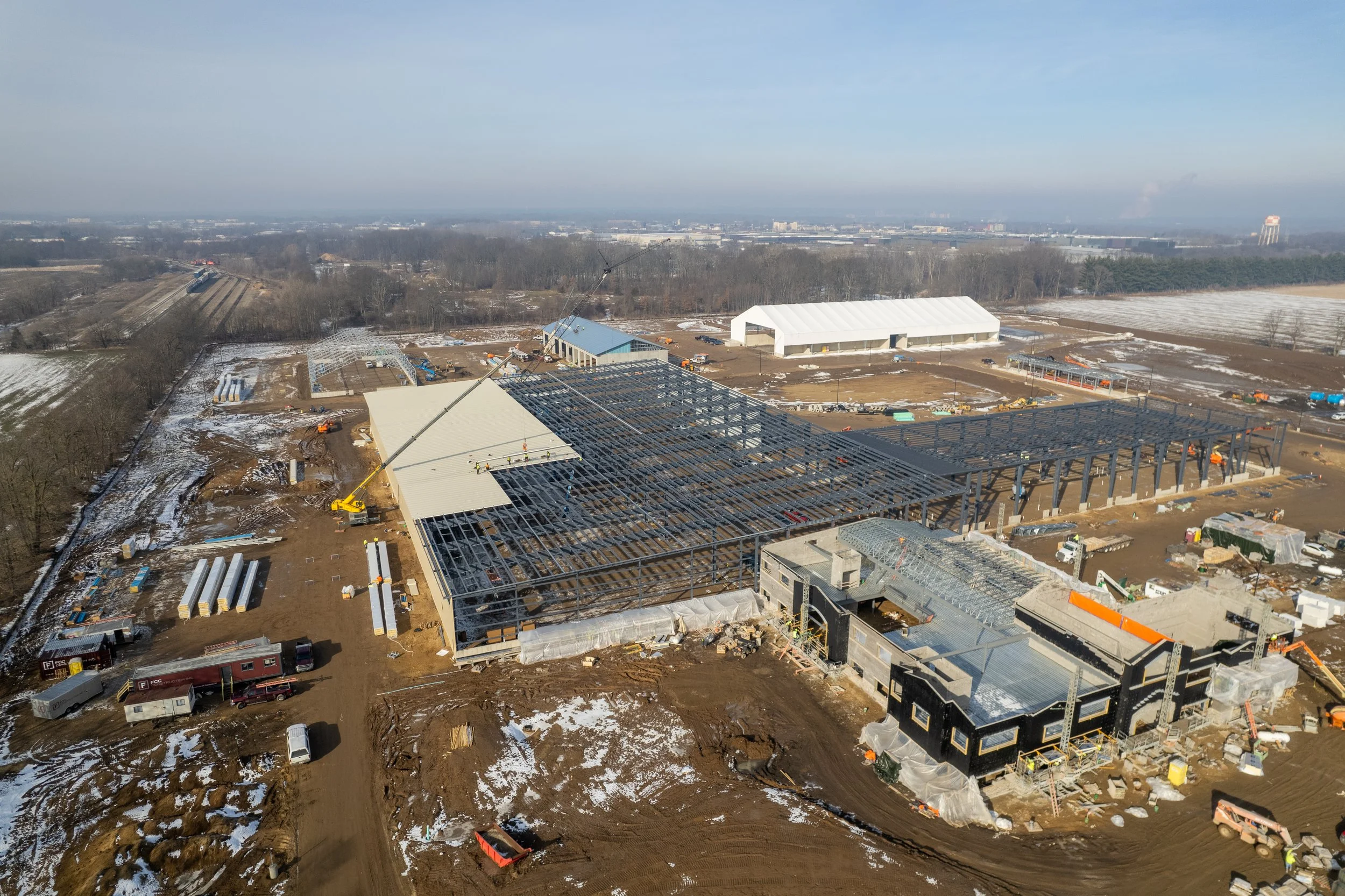 Aerial view of a large construction site with steel framework for a building, multiple construction vehicles, and snow patches on the ground.