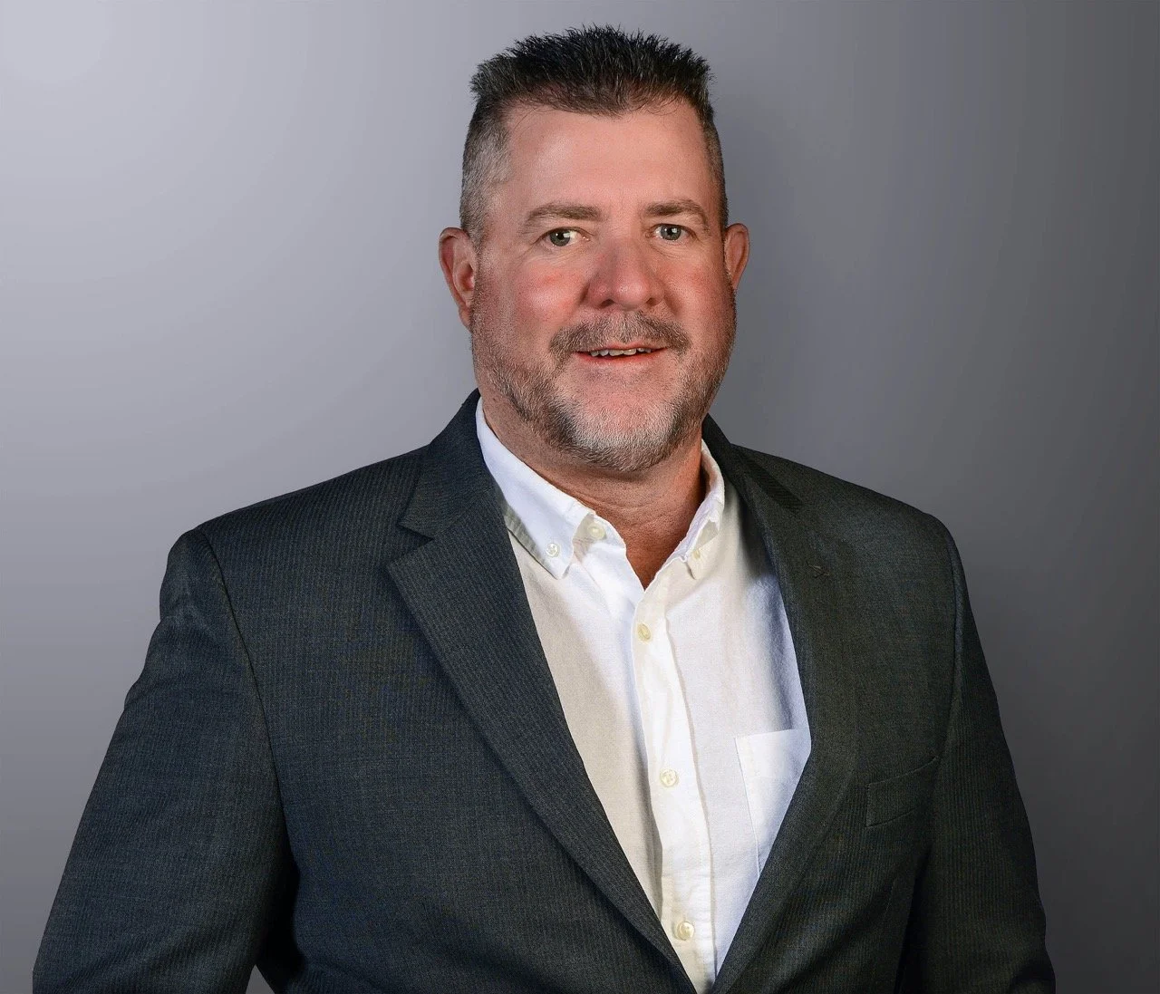 A professional portrait of a middle-aged man with short dark hair and a beard, wearing a black suit jacket and white button-down shirt, smiling against a gray background.