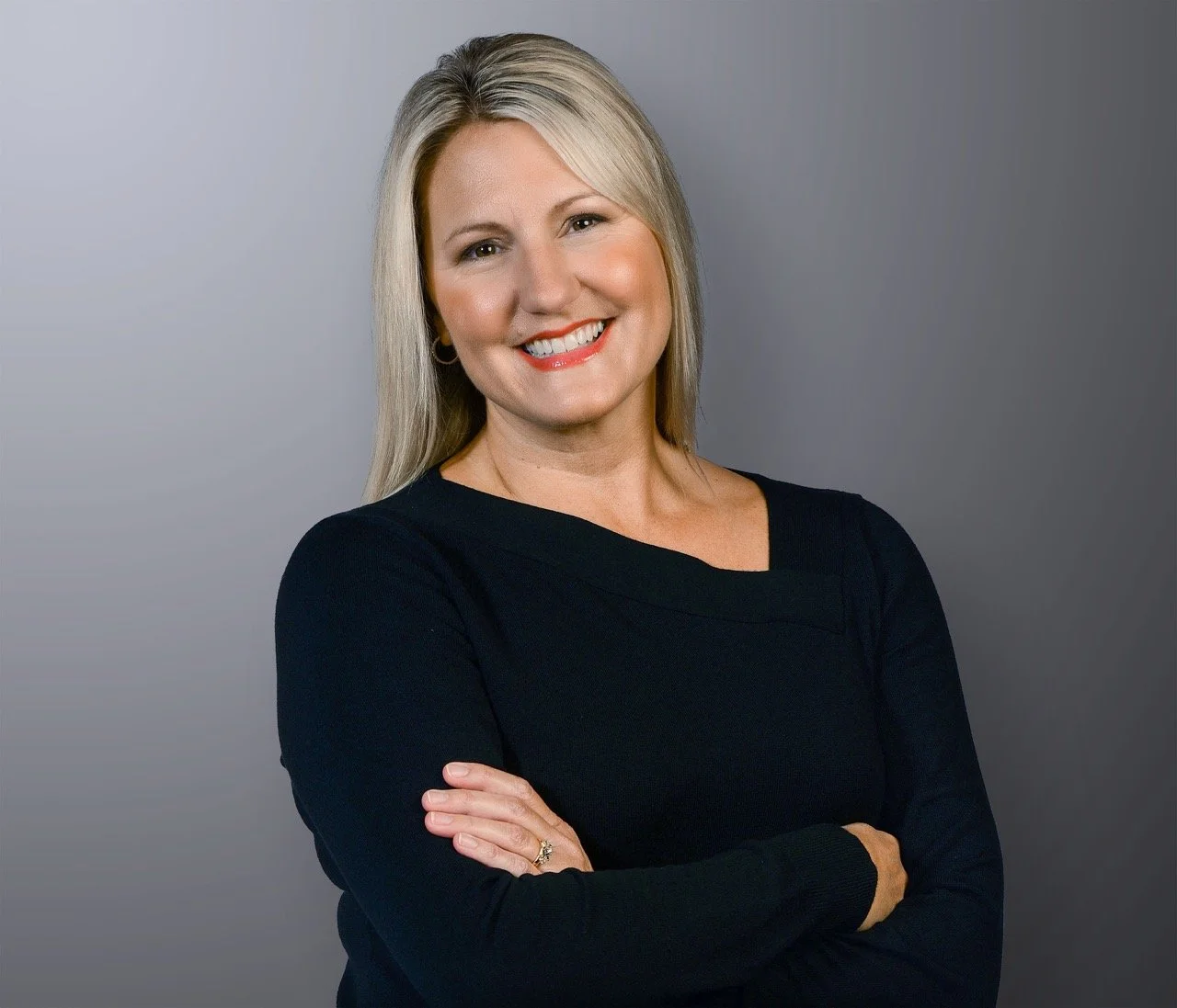 A portrait of a smiling woman with blonde hair, wearing a black top, arms crossed, standing against a gray background.