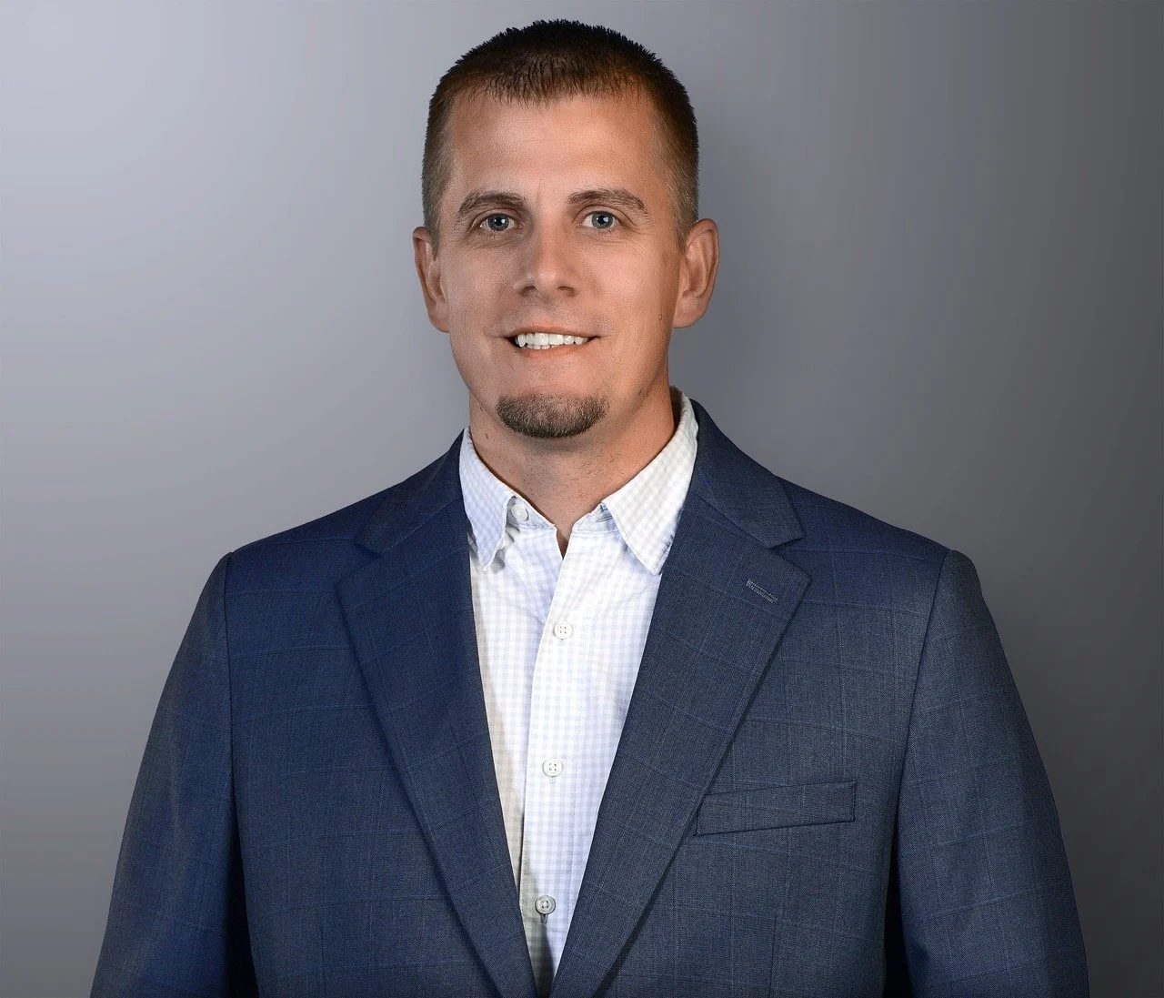 Headshot of a smiling man with short brown hair and a goatee, wearing a dark blue suit jacket and a white button-up shirt, standing against a gray background.