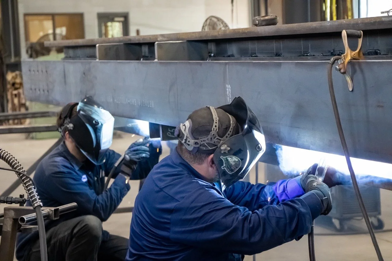Two workers welding a large metal beam in an industrial workshop.