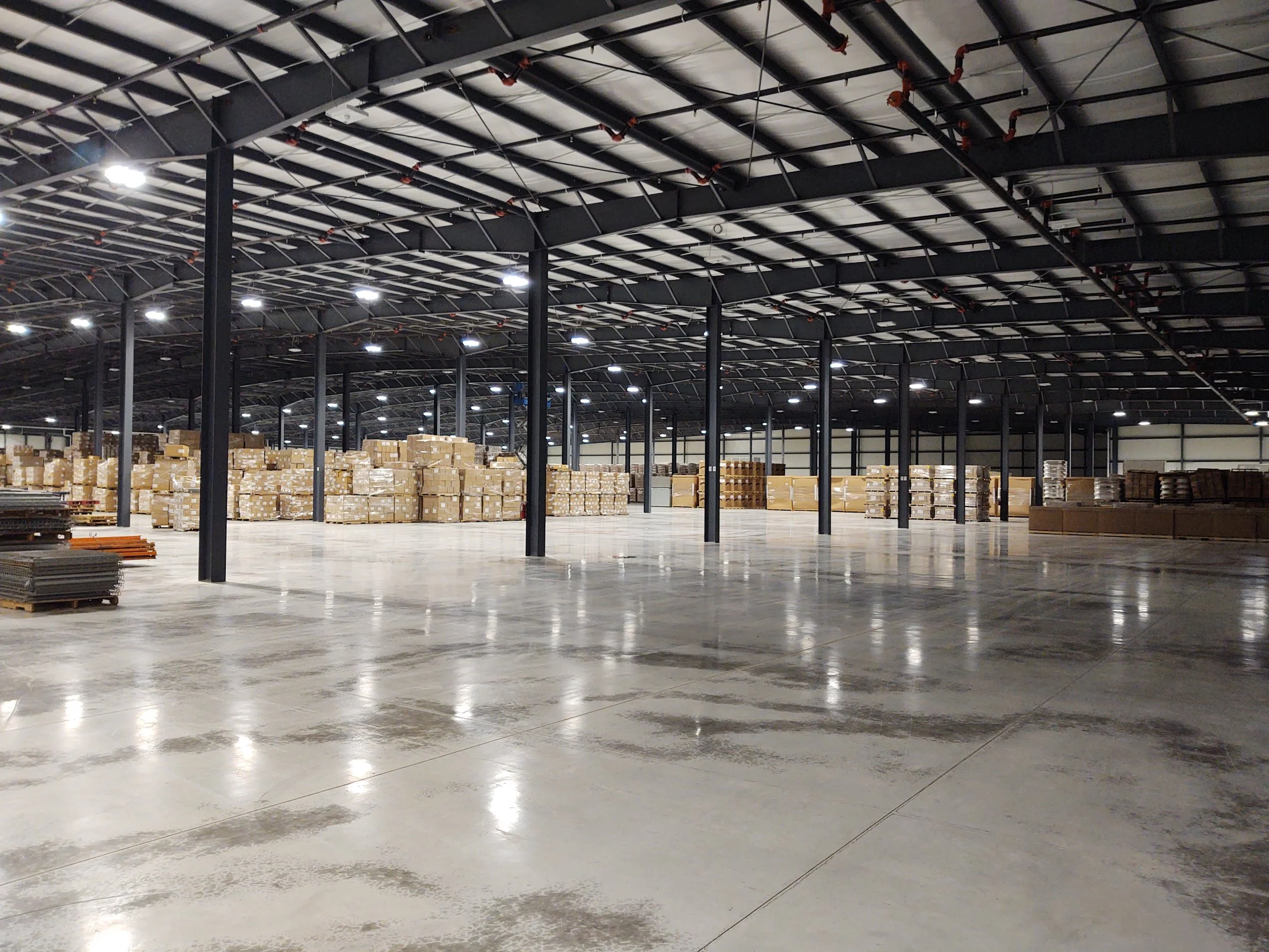 Empty warehouse with stacked boxes and pallets on the left side, metal support beams, and bright ceiling lights.
