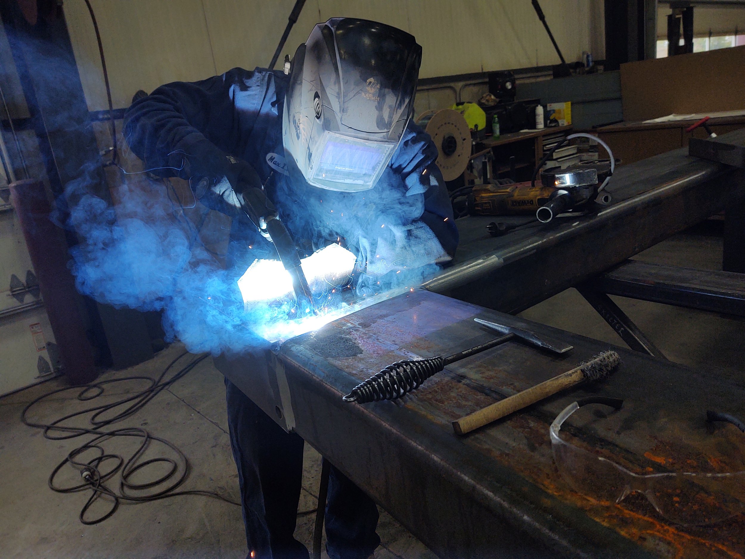 A person welding metal at a workbench in a workshop, wearing a protective welding helmet and gloves, with sparks and smoke around the welding area.