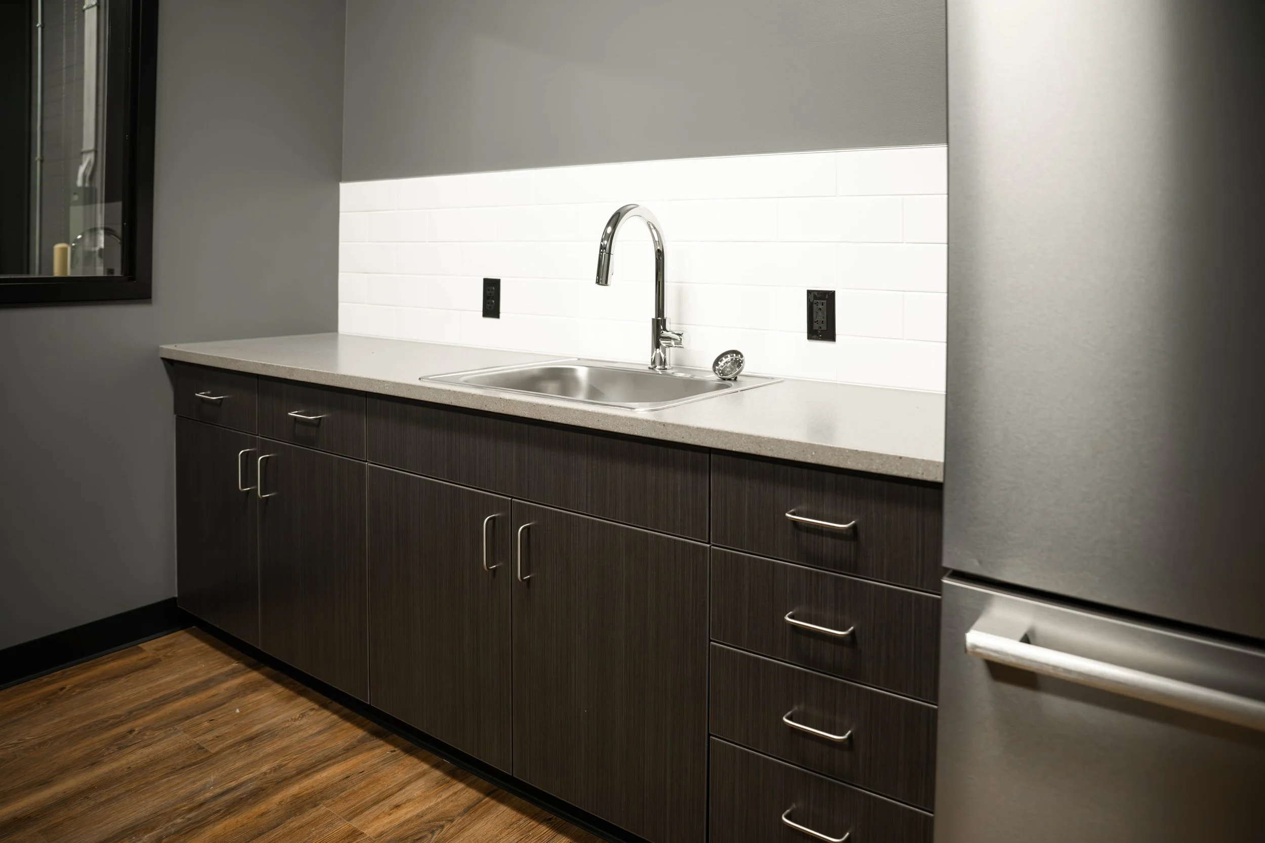 Breakroom kitchenette with a stainless sink, dark cabinetry and a white tile backsplash at Weller Auto