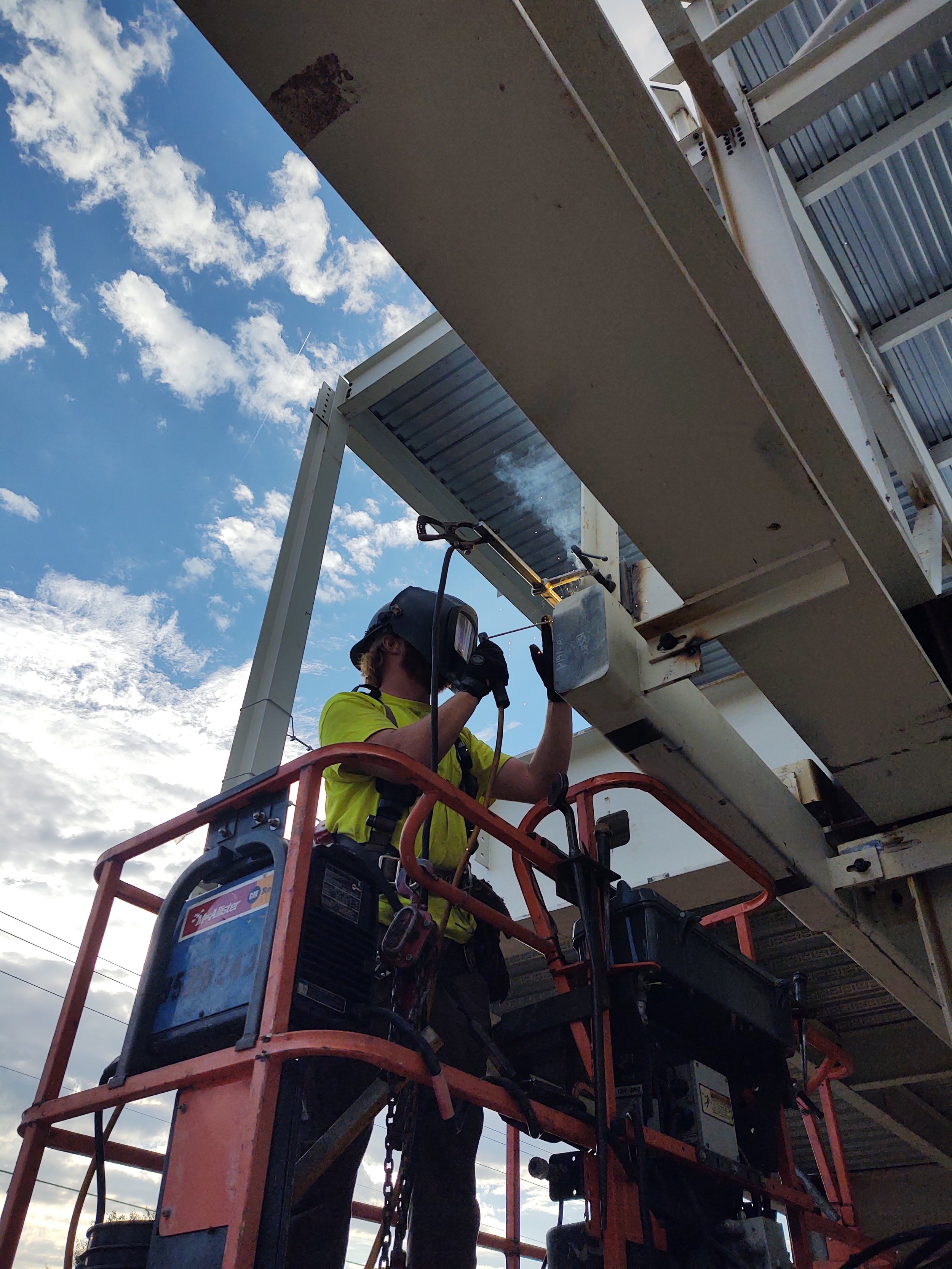 A worker wearing a yellow shirt, safety helmet, and gloves welding a metal structure on an orange lift platform against a background of a partly cloudy sky.