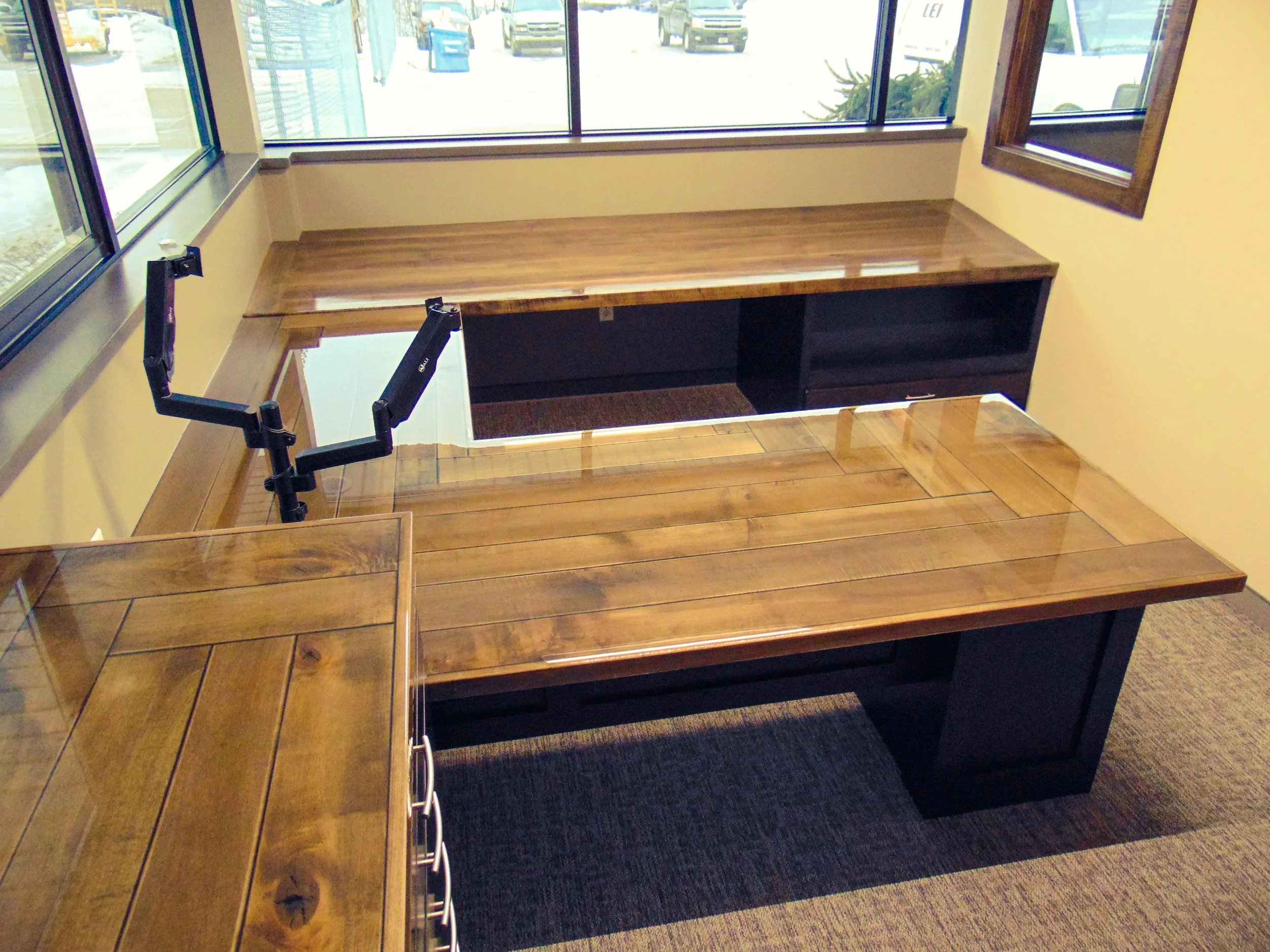 Interior of an office with a wooden L-shaped desk, a black shelving unit, and large windows showing parked cars outside.