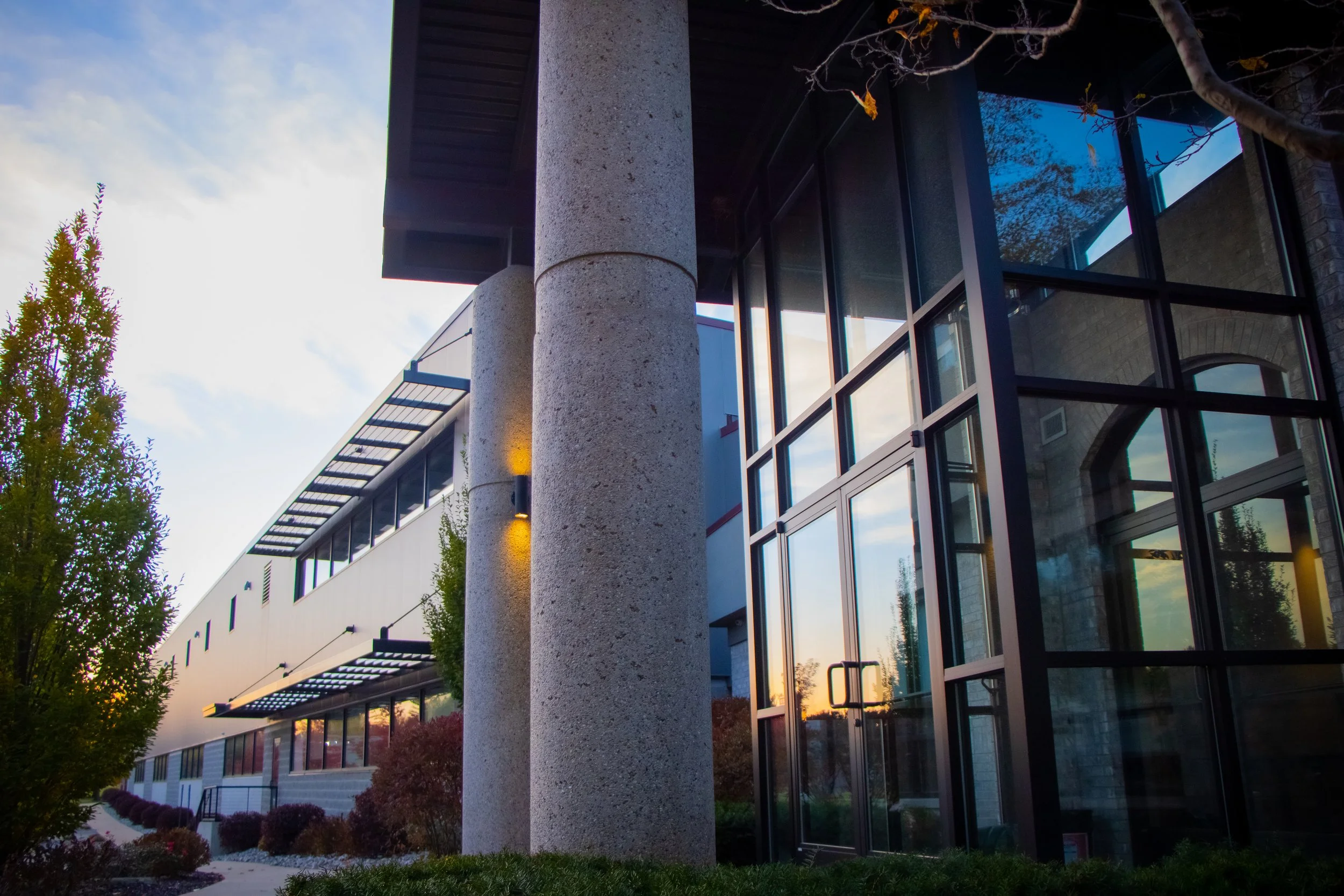 Modern building with large glass windows, concrete pillars, and outdoor lighting, surrounded by trees and landscaping at sunset.