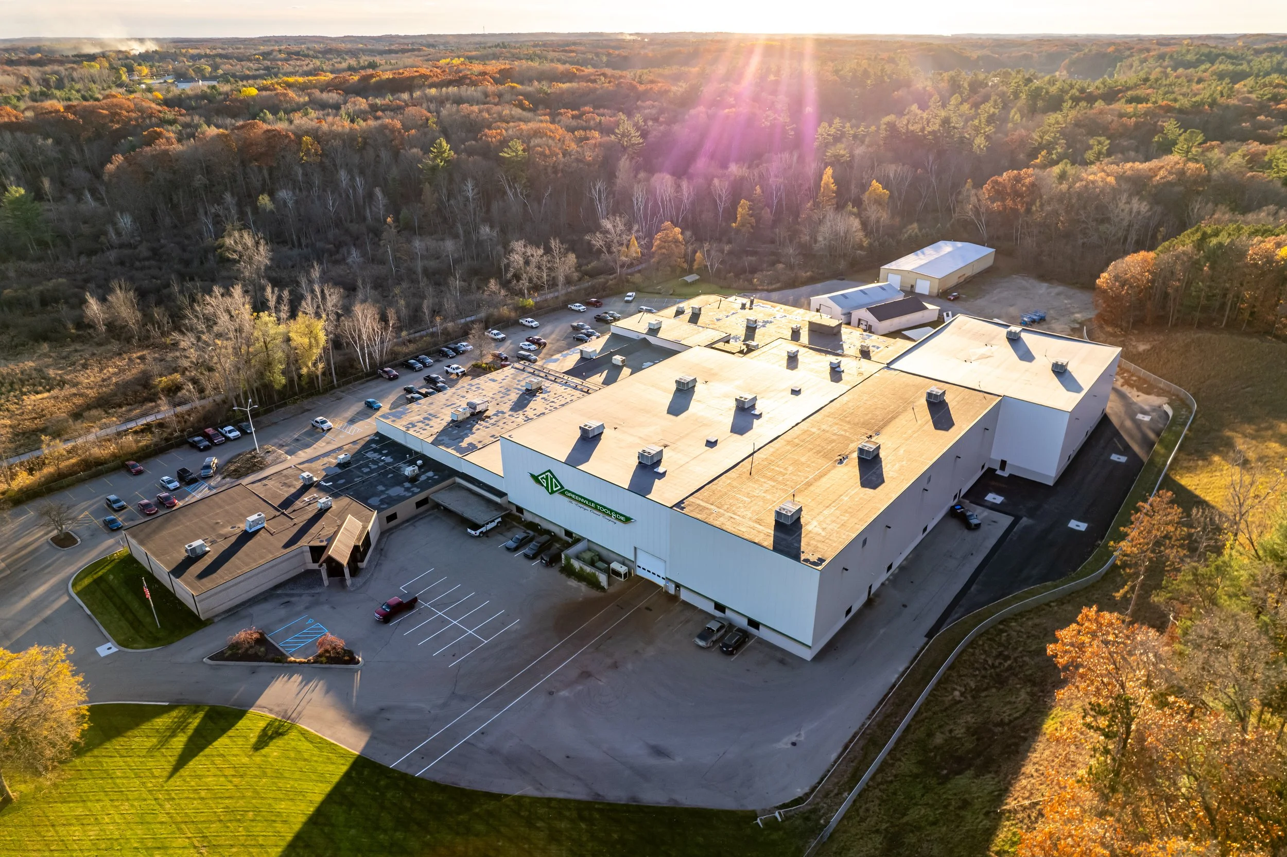 Aerial view of a large building with a parking lot, surrounded by trees with autumn foliage, and a forest in the background, during sunset.