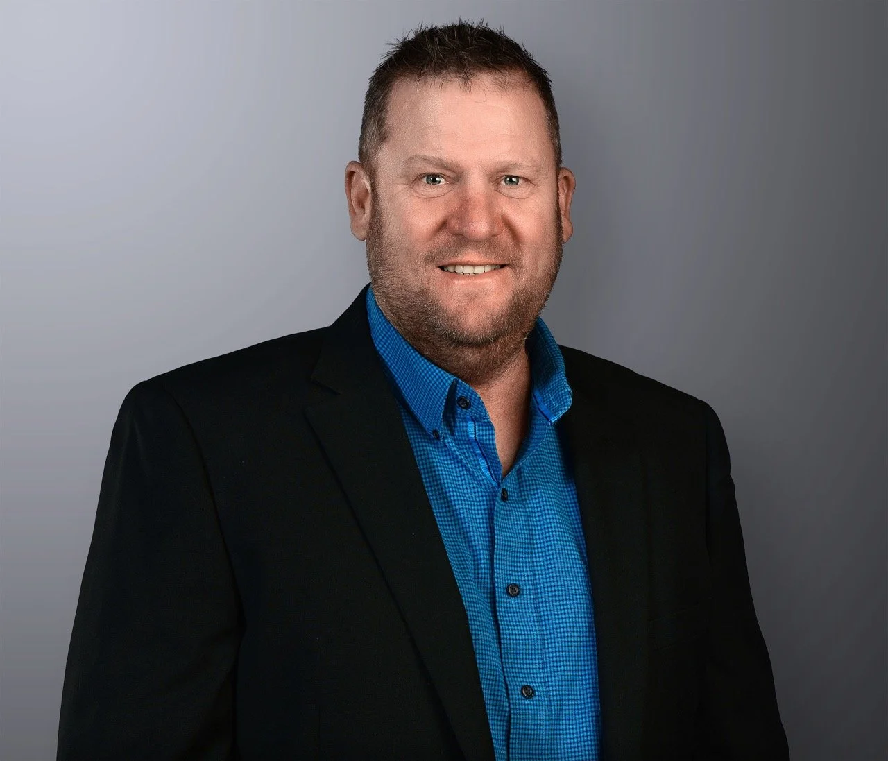A professional headshot of a man with short brown hair, a beard, wearing a blue shirt and a black blazer, smiling against a gray background.