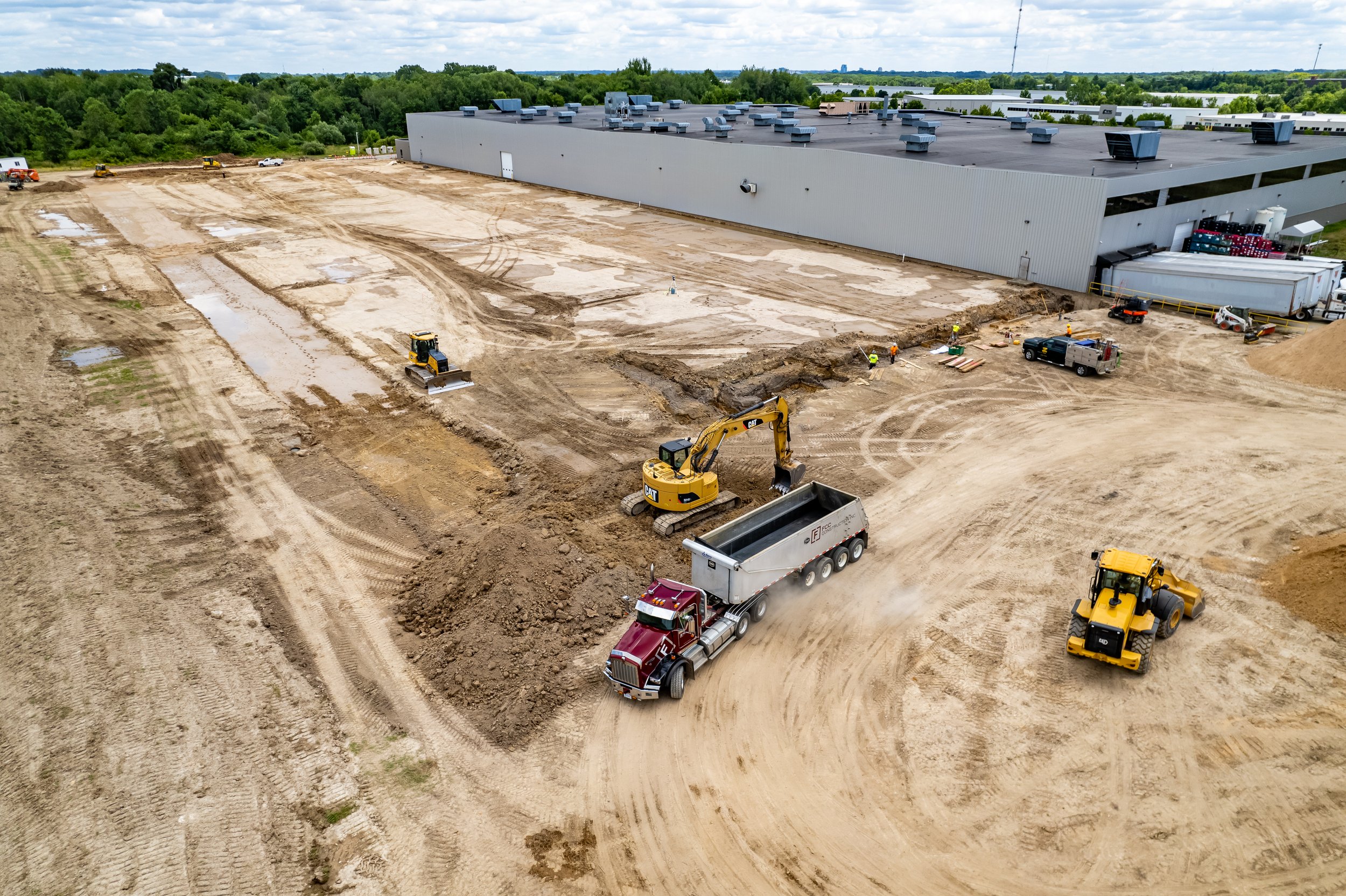 Construction site with heavy machinery, including a bulldozer, excavator, and dump truck, leveling and moving dirt near a large industrial building.