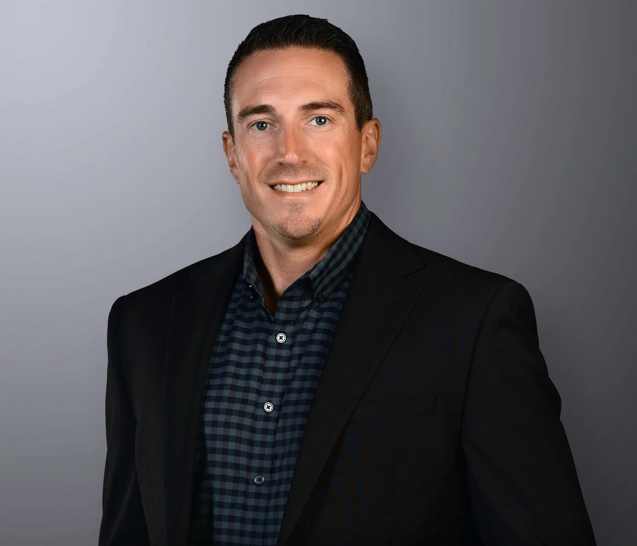 Professional headshot of a man with short dark hair, blue eyes, wearing a dark blazer and checkered shirt, smiling against a gray background.