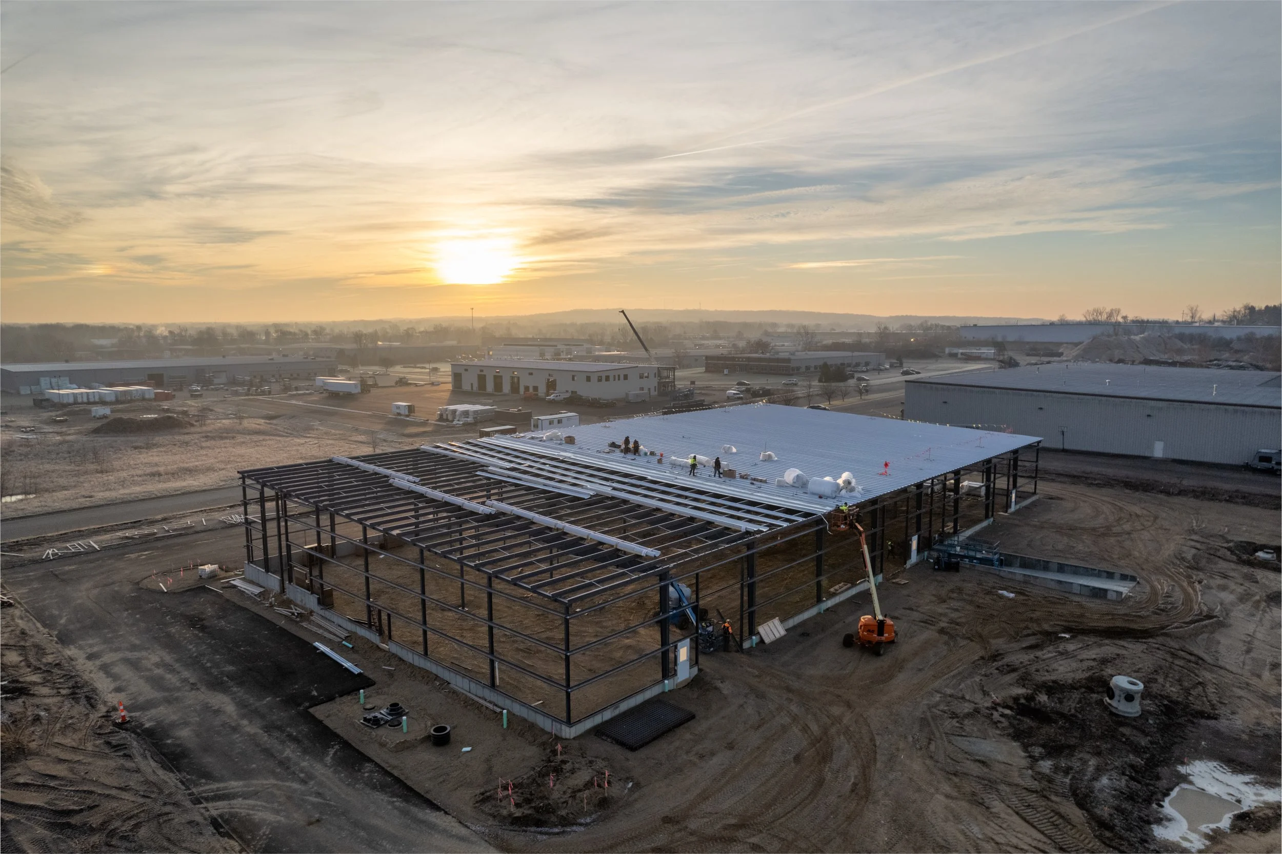 Construction site with steel framework and roof installation ongoing, and workers on the site during sunset.