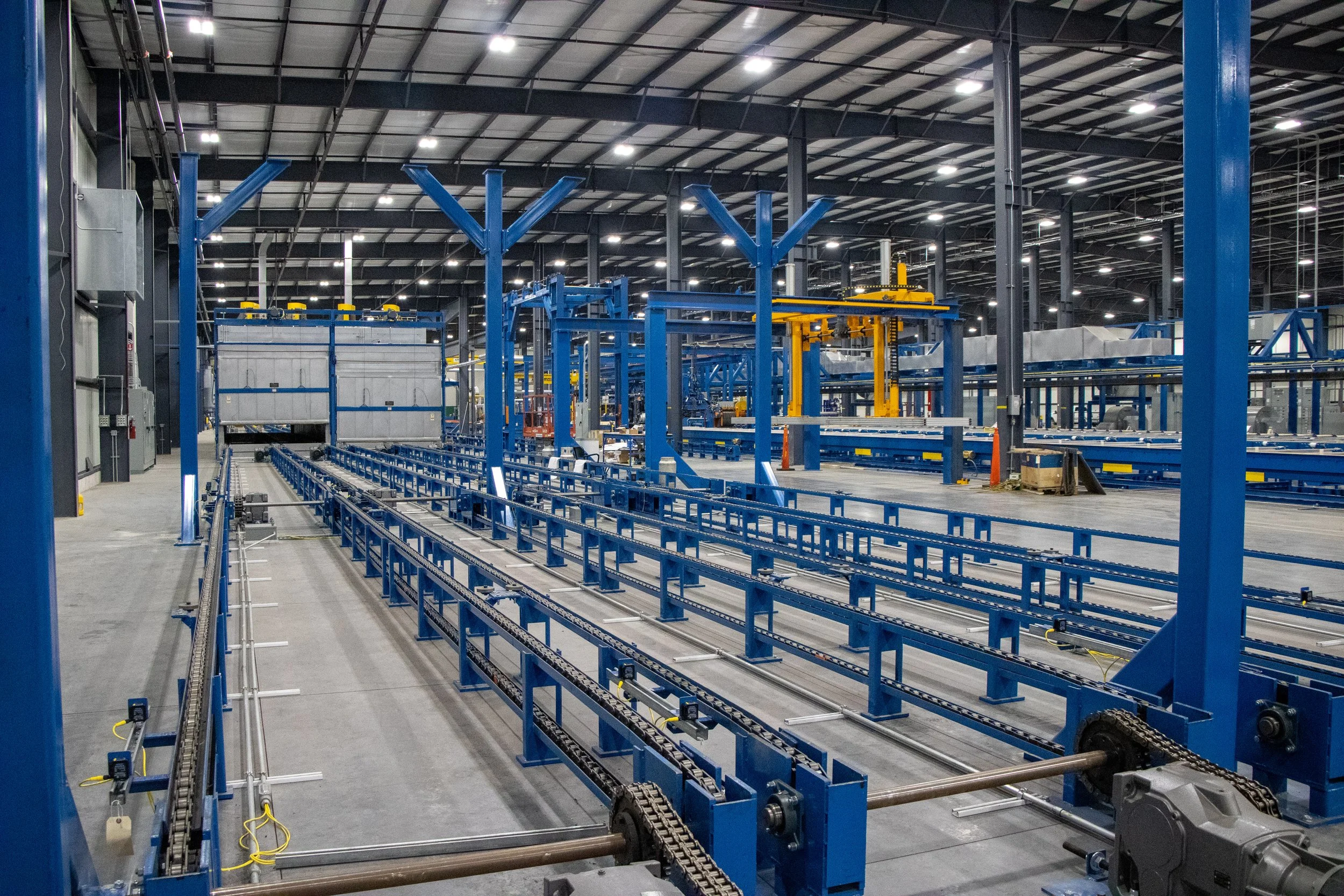 Interior view of an empty industrial warehouse or factory with blue metal framework, conveyor tracks, and overhead equipment, illuminated by ceiling lights.