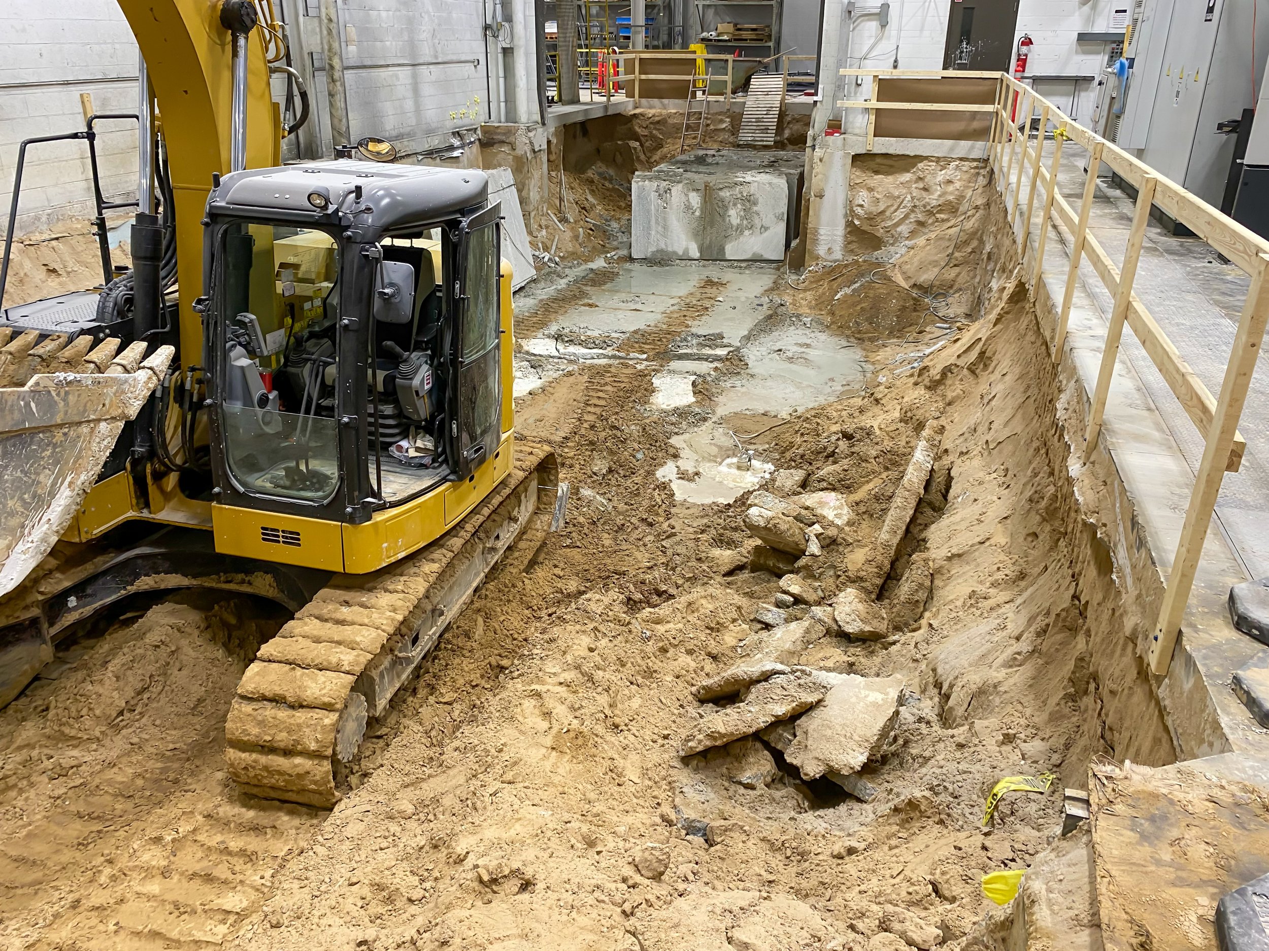 Construction site with excavator digging a trench inside an industrial building. There are wooden safety railings along the edge of the trench.
