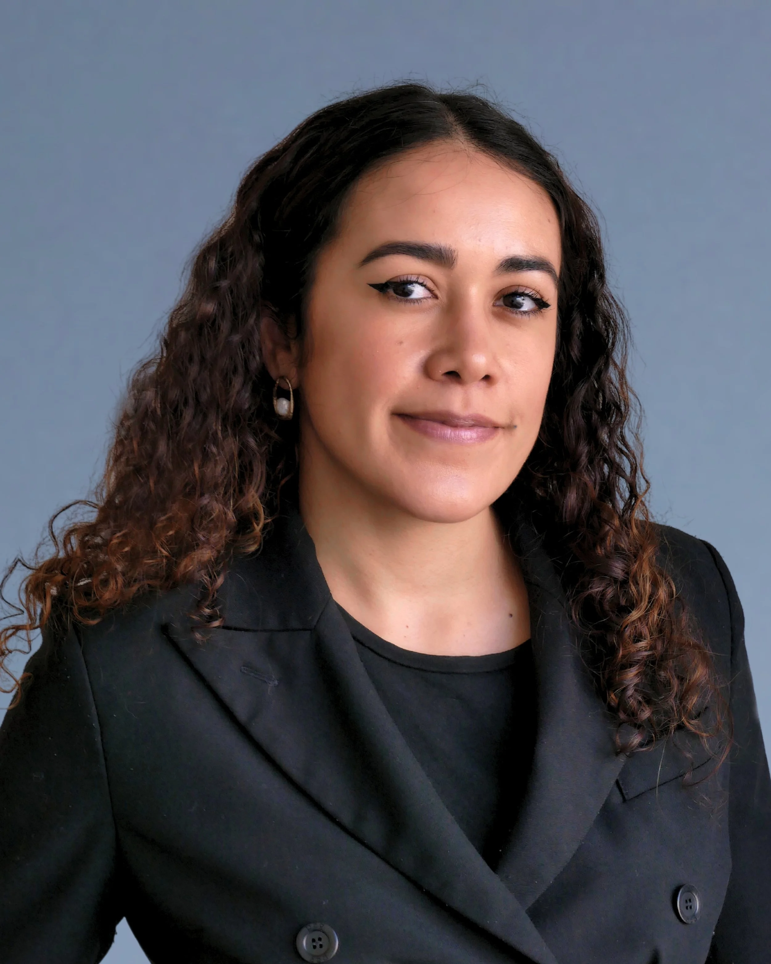 A young woman with curly dark hair wearing a black blazer, pearl earrings, and makeup, posing against a plain gray background.