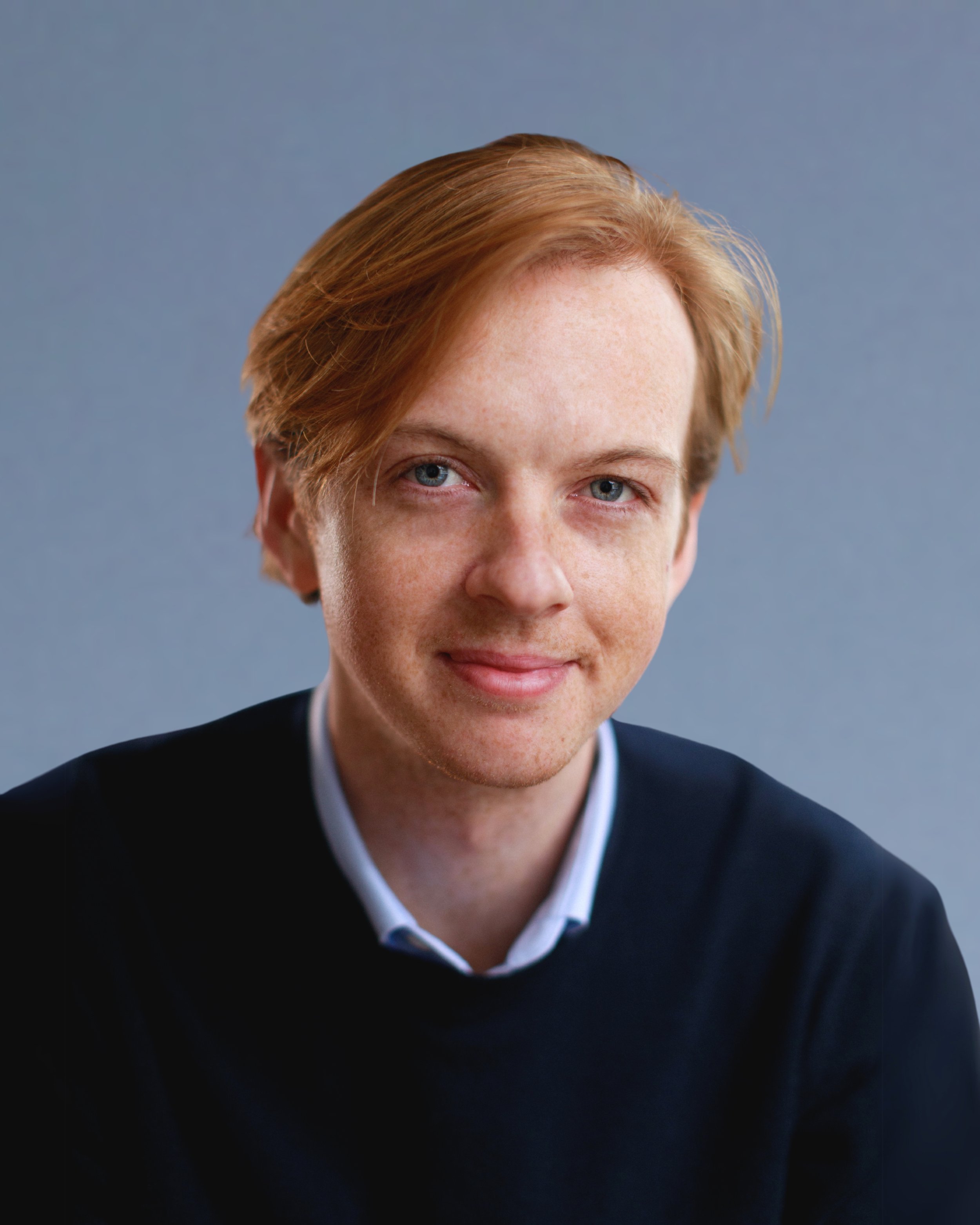A young man with red hair, blue eyes, and freckles, wearing a dark sweater over a collared shirt, smiling at the camera against a plain gray background.