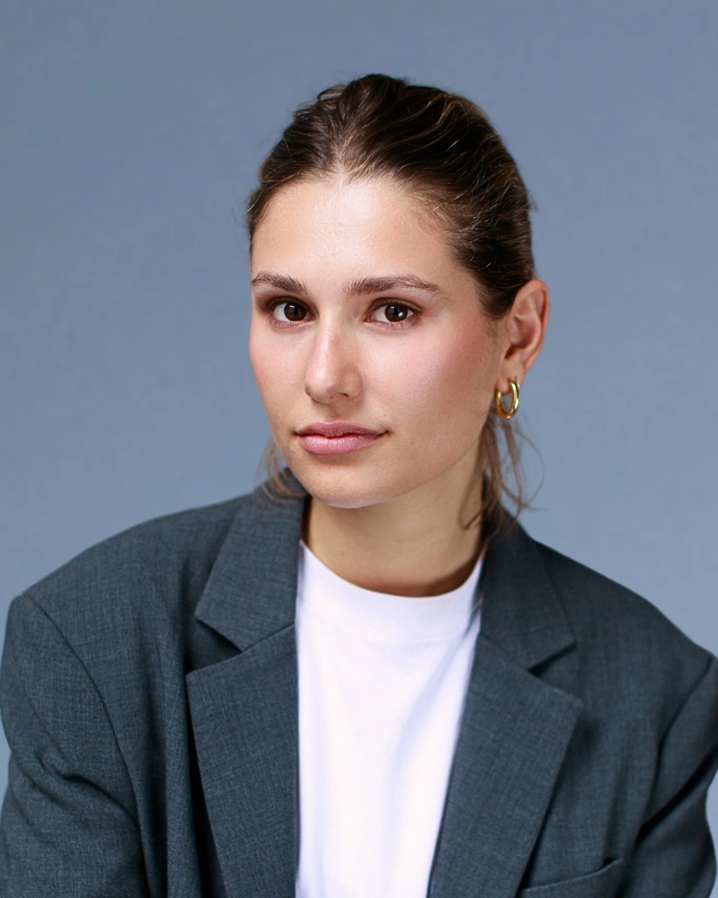 A woman with brown hair tied back, wearing a dark blazer, white shirt, and gold hoop earrings, looking at the camera against a blue background.