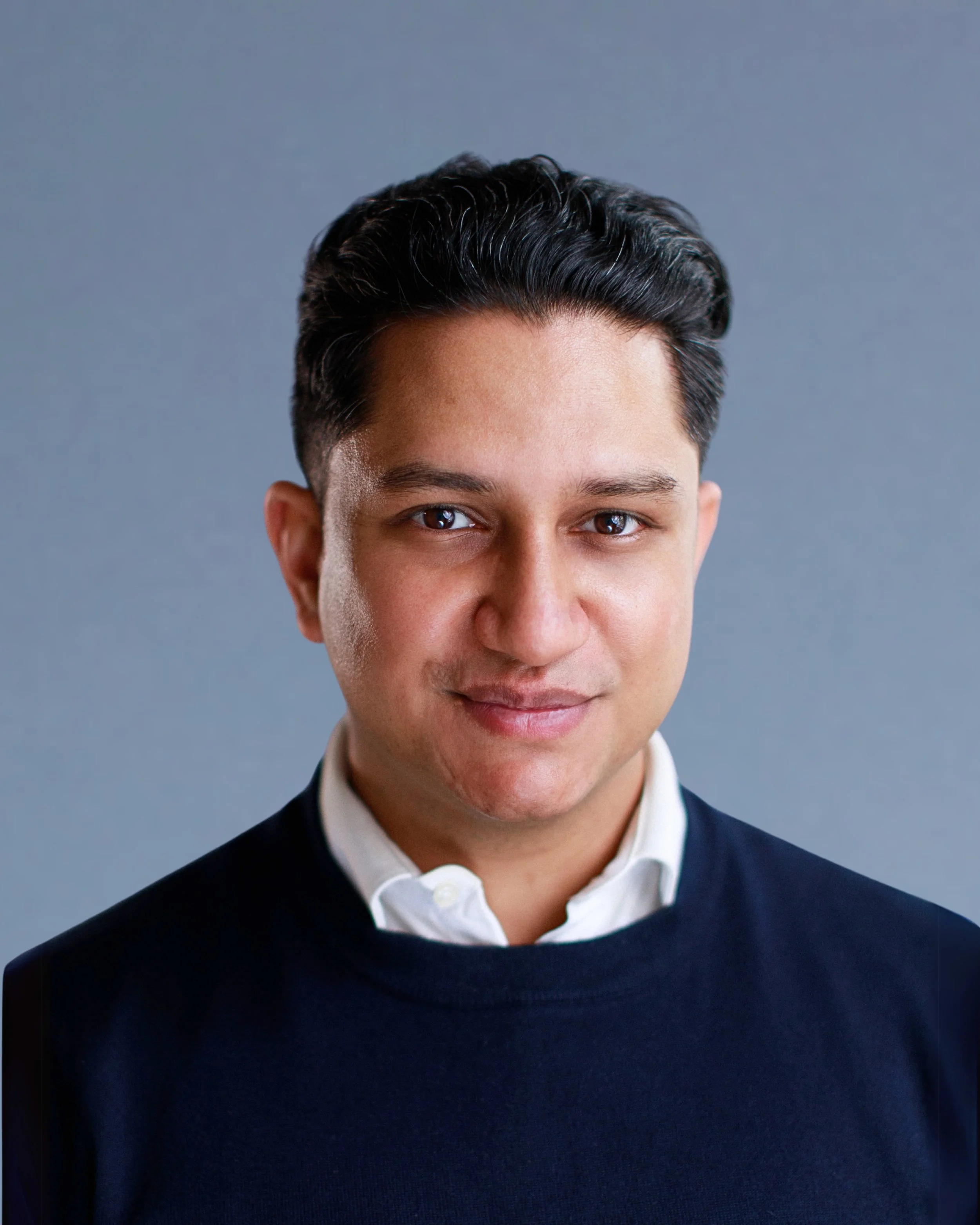 Close-up portrait of a young man with dark hair, wearing a white shirt and navy sweater, smiling gently against a neutral background.