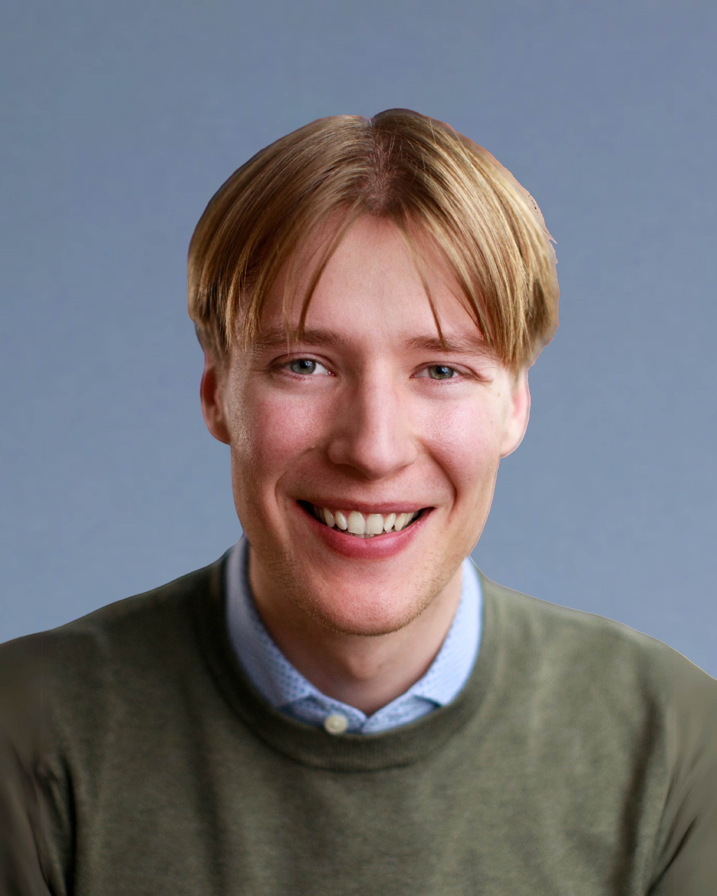 A young man with reddish-brown hair and blue eyes smiling, wearing a green sweater over a collared shirt, against a plain blue background.