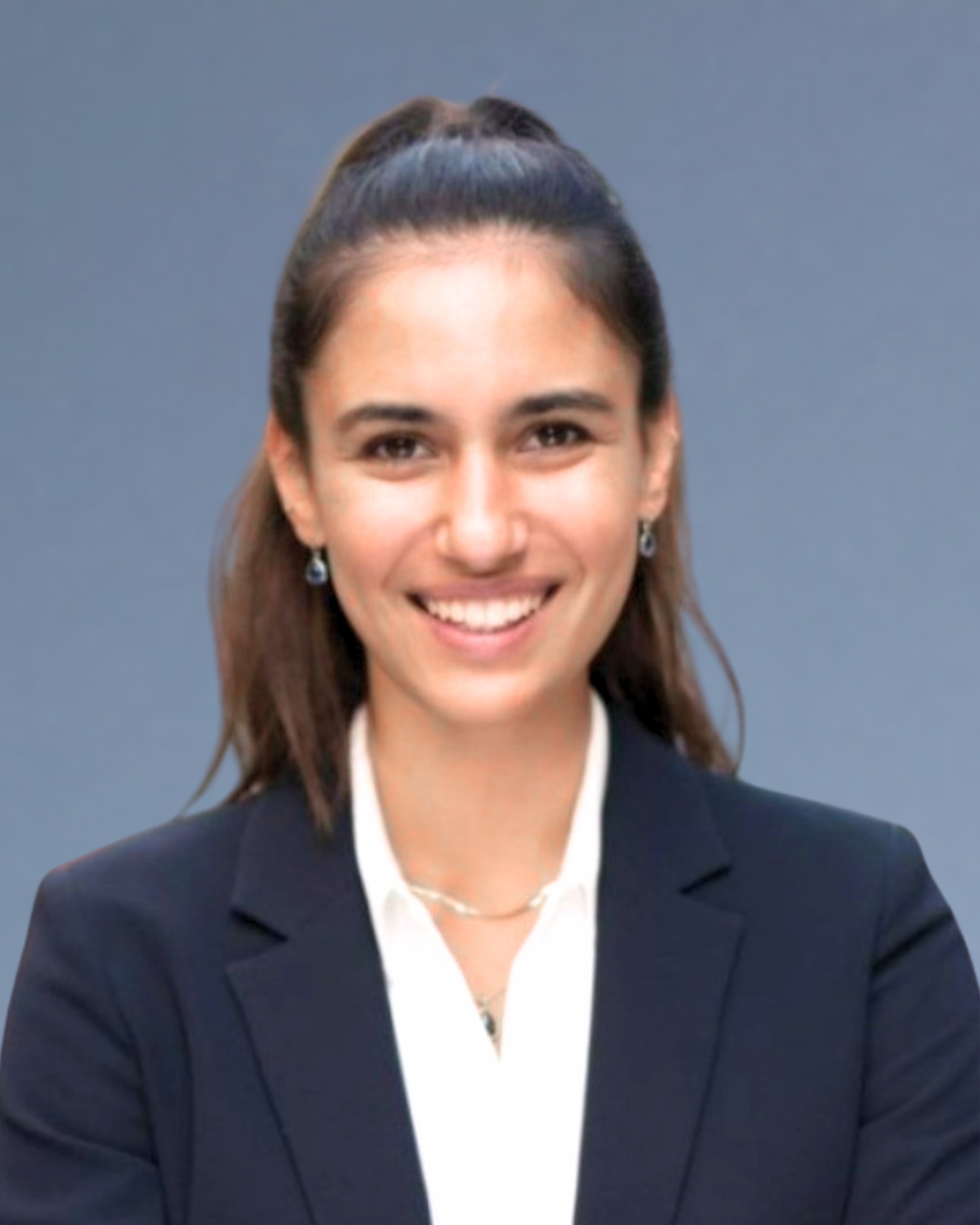 A professional woman smiling, wearing a black blazer and white shirt, with earrings and a necklace, against a gray background.