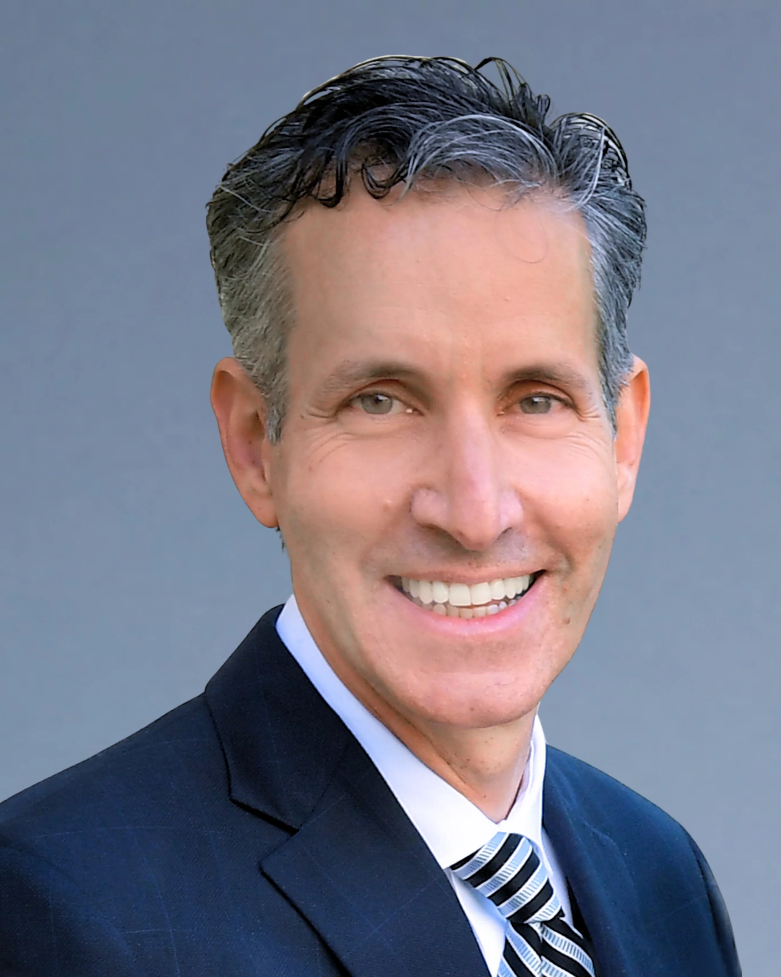A smiling man in a dark suit with a striped tie, with gray hair and a light skin tone, against a plain gray background.